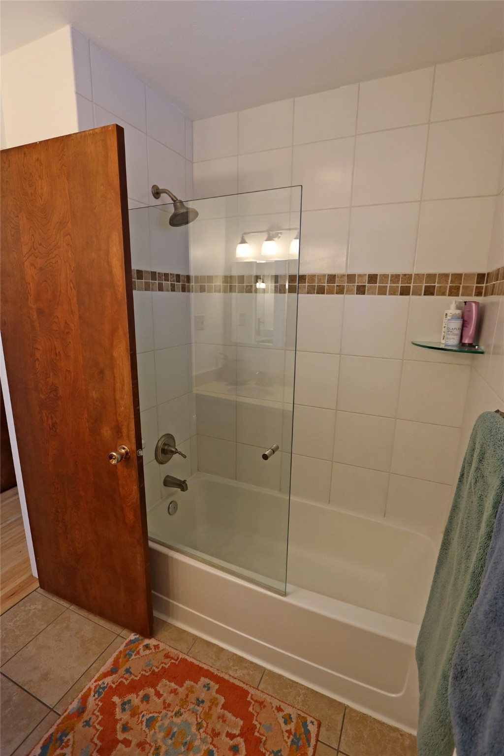 Bathroom with a glass-screened bathtub, white tile, and brown mosaic accent strip.