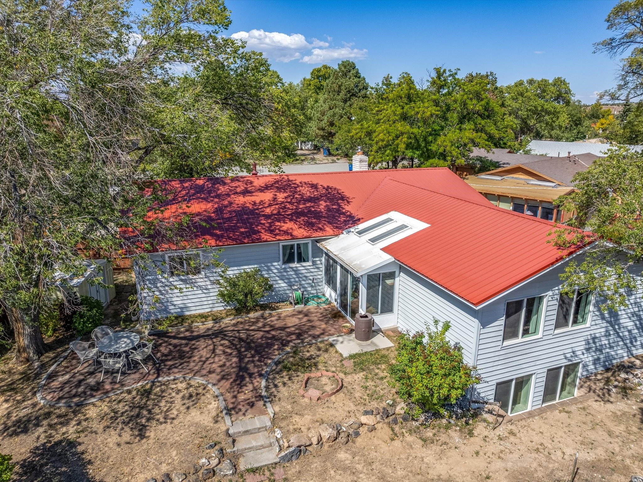 Aerial view of a gray home with a red metal roof, skylights, and a tree-shaded patio.