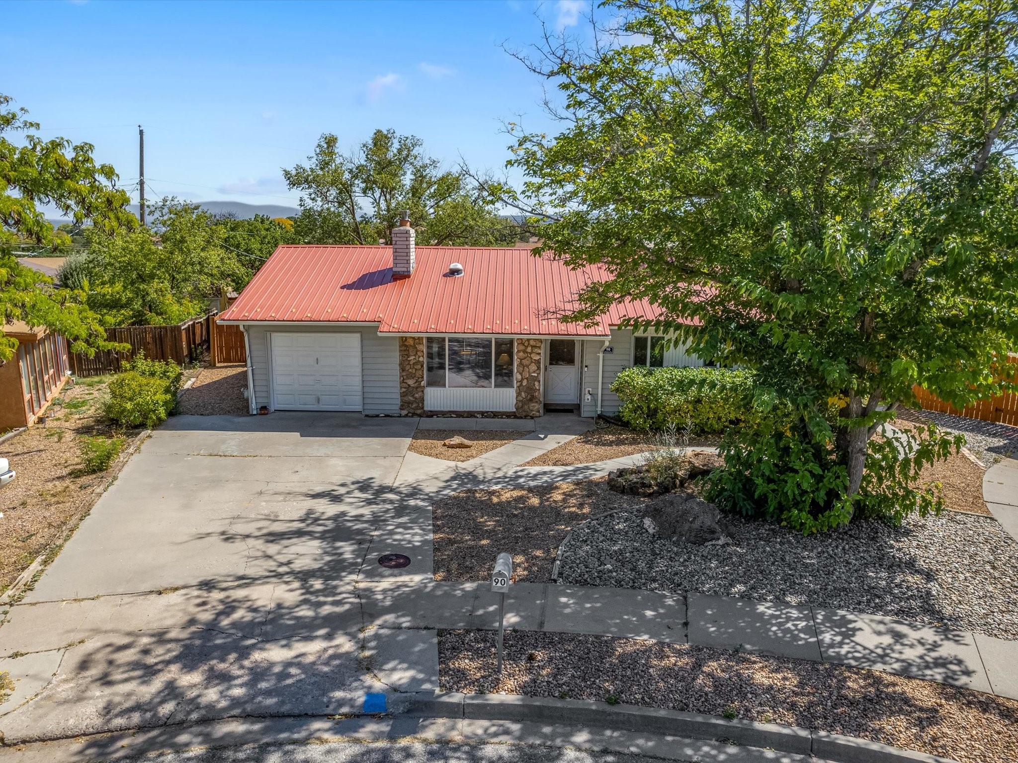 Single-story home with a red metal roof, stone facade accents, attached garage, and mature shade trees.