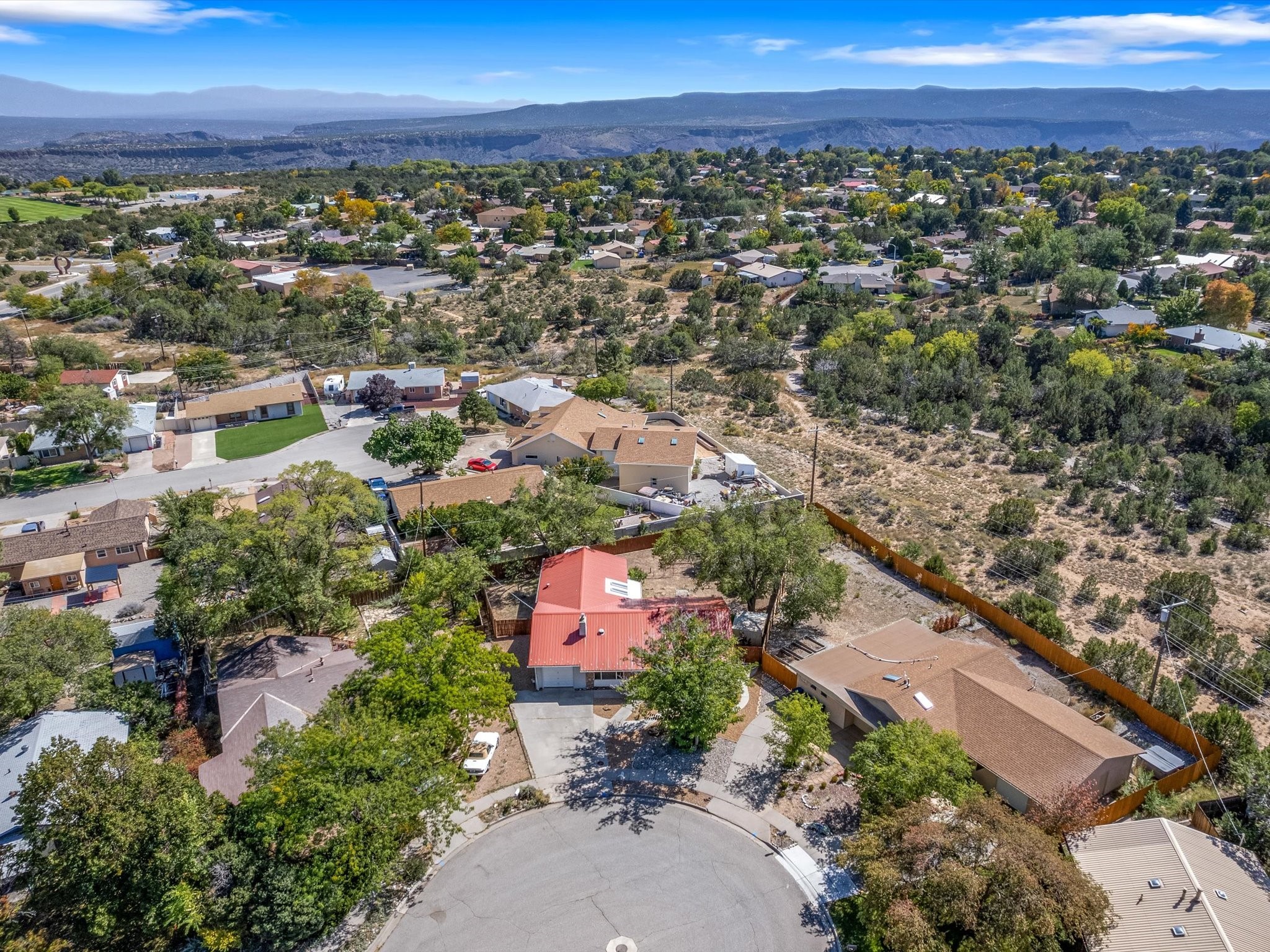 Aerial view of a red-roofed home in a tree-lined neighborhood, with dry open land and distant mountains.
