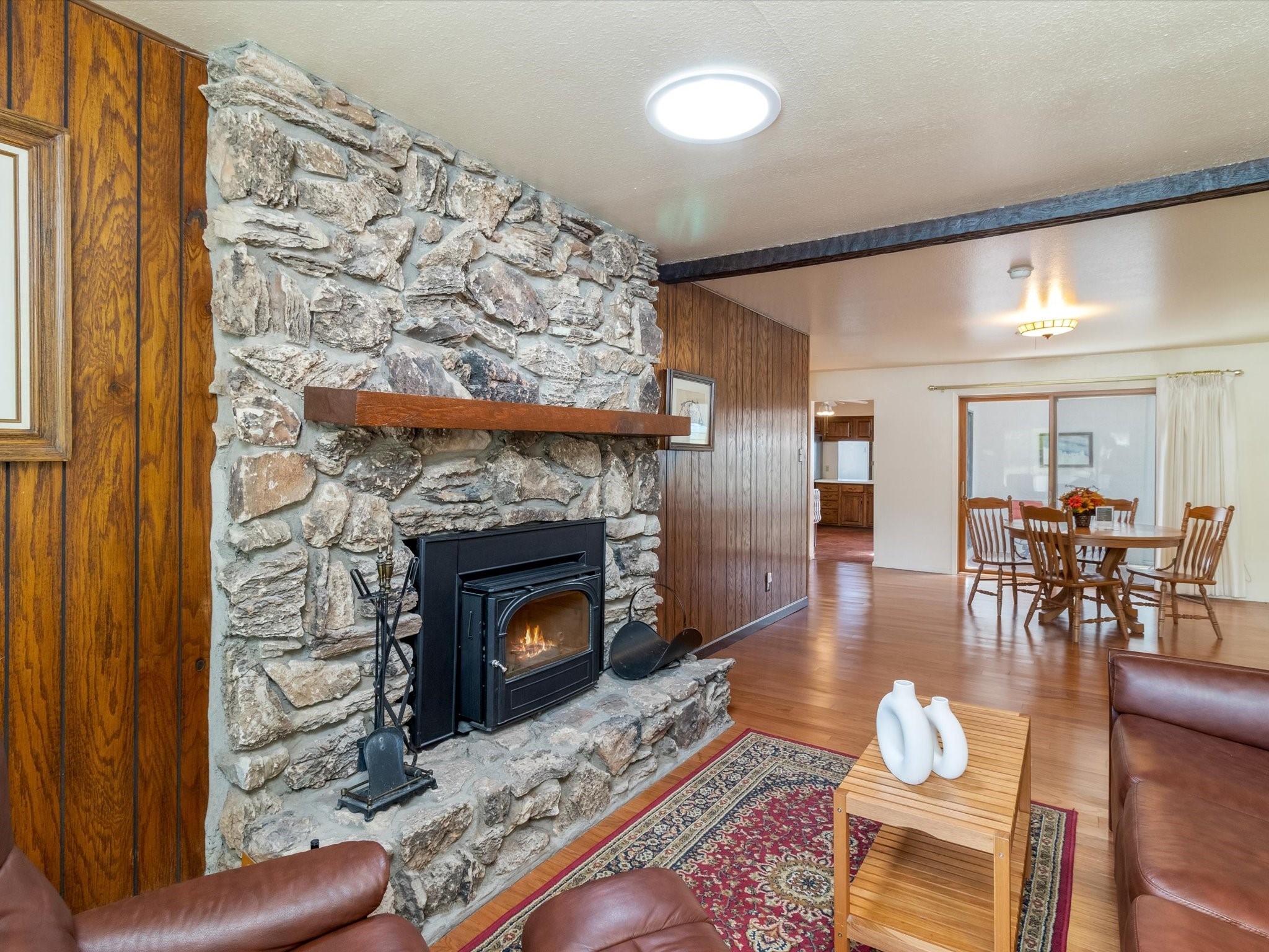 Living room with a stone fireplace and wood stove beside a dining area with sliding glass doors.