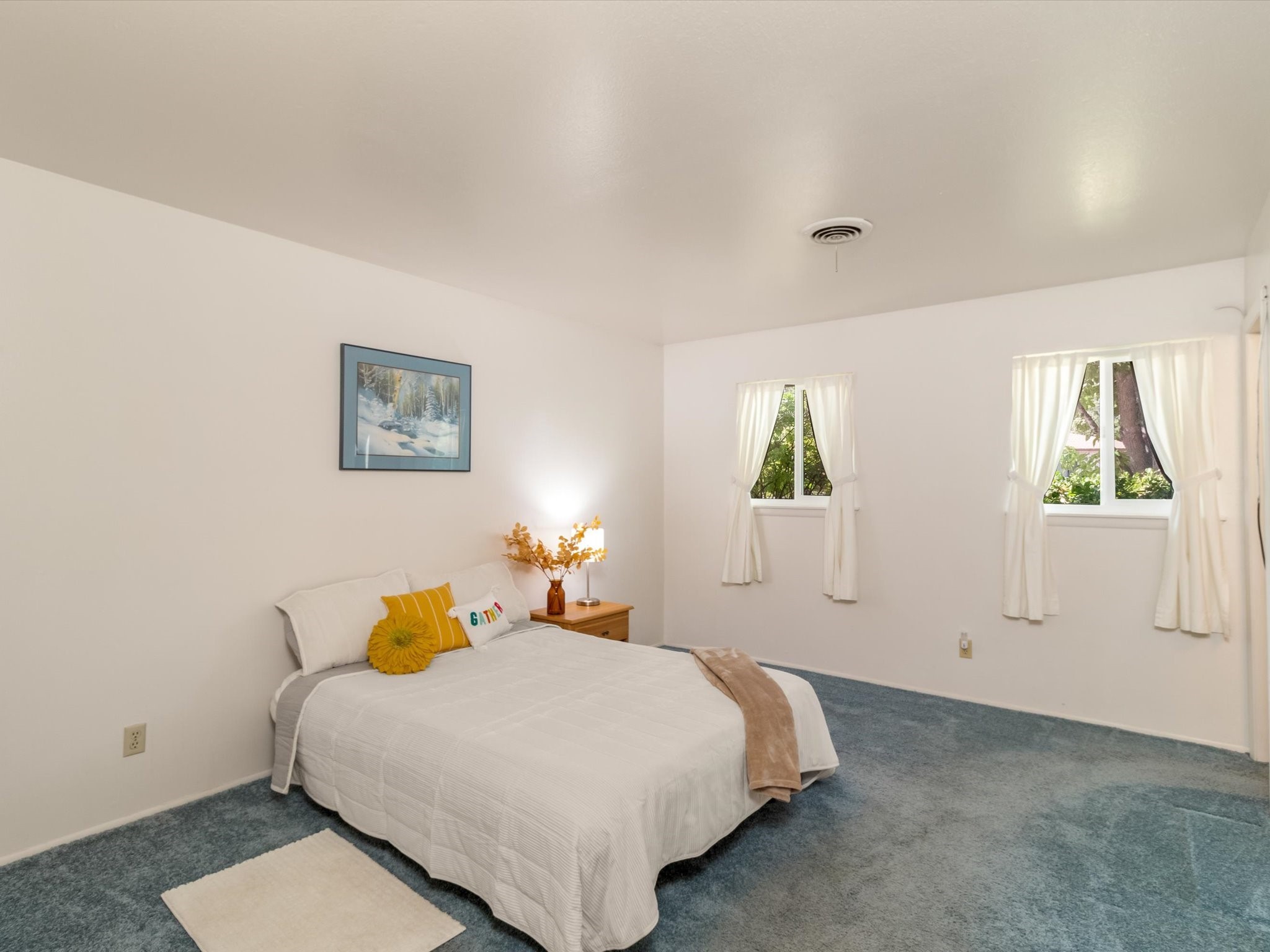 Furnished bedroom with a white bed, blue-gray carpet, and two curtained windows.