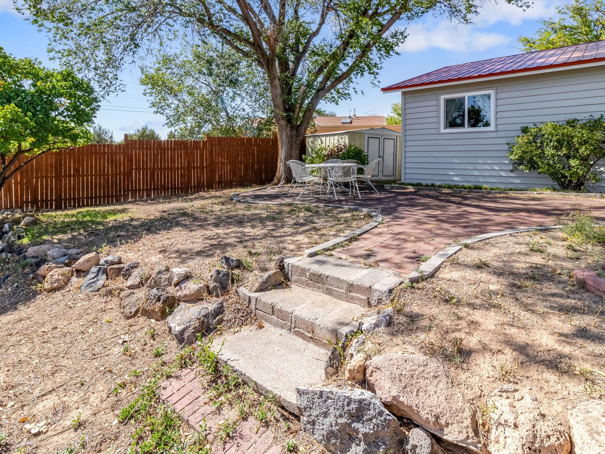 Backyard patio with a red paver walkway, rock-edged steps, metal seating, and a large shade tree.