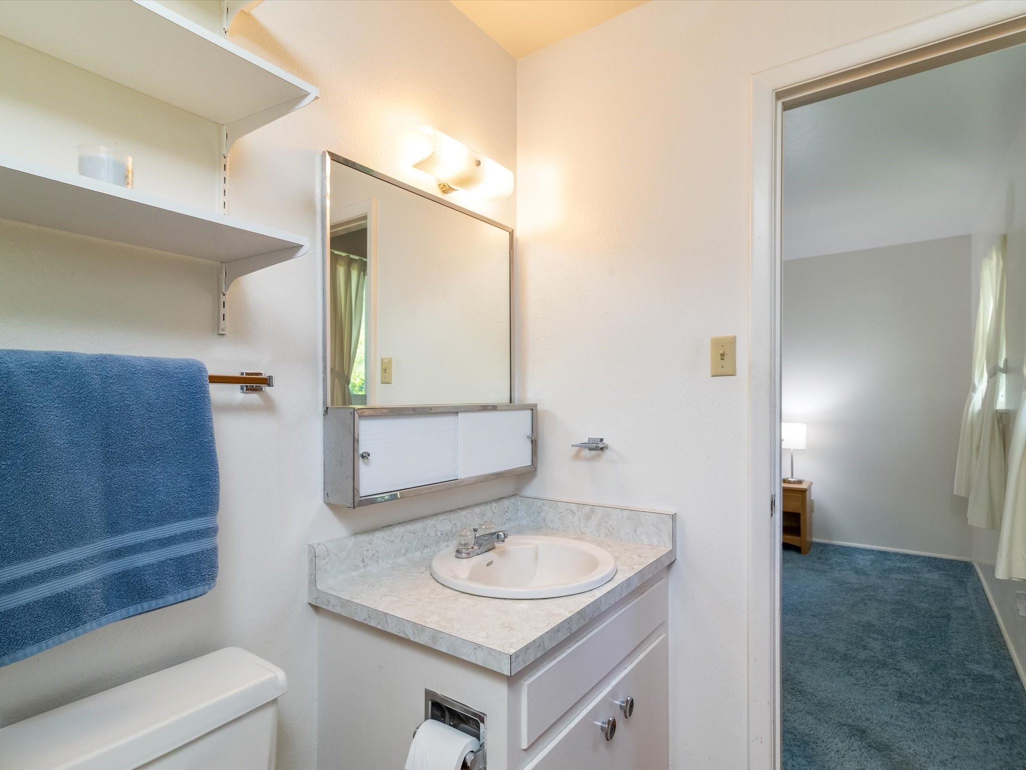 Bathroom vanity with a mirrored medicine cabinet, open shelves, and a blue towel beside the doorway.