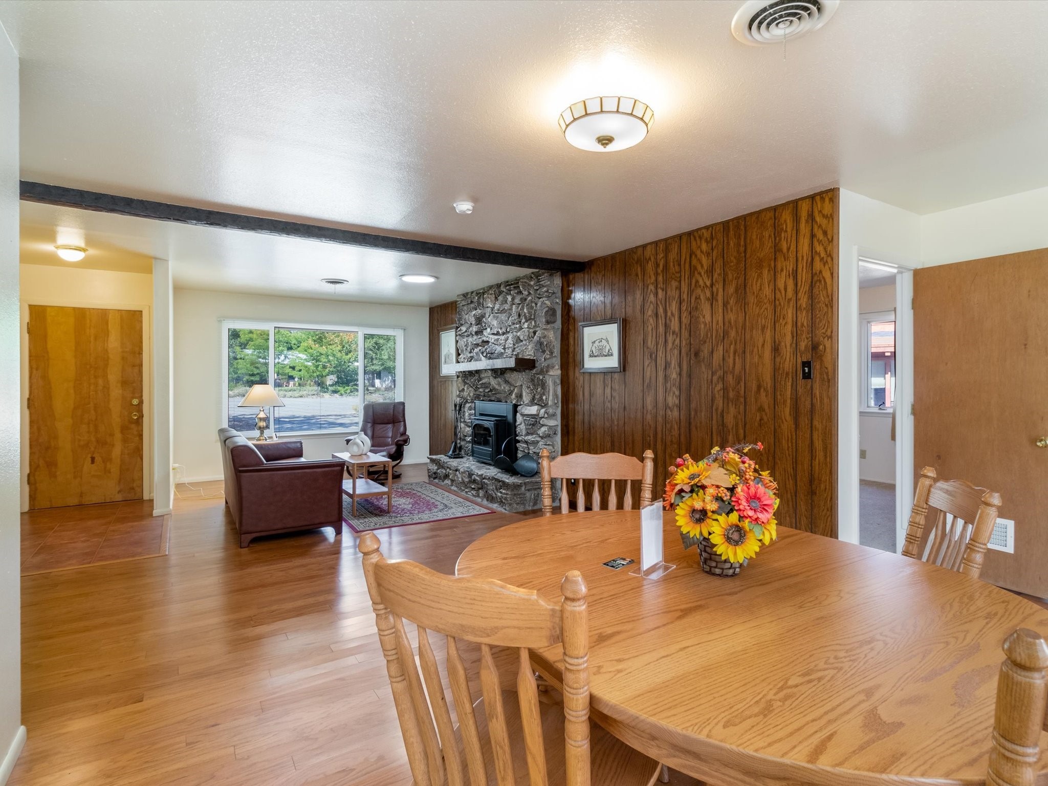 Dining and living area with a wooden table, stone fireplace, and wood-paneled wall.