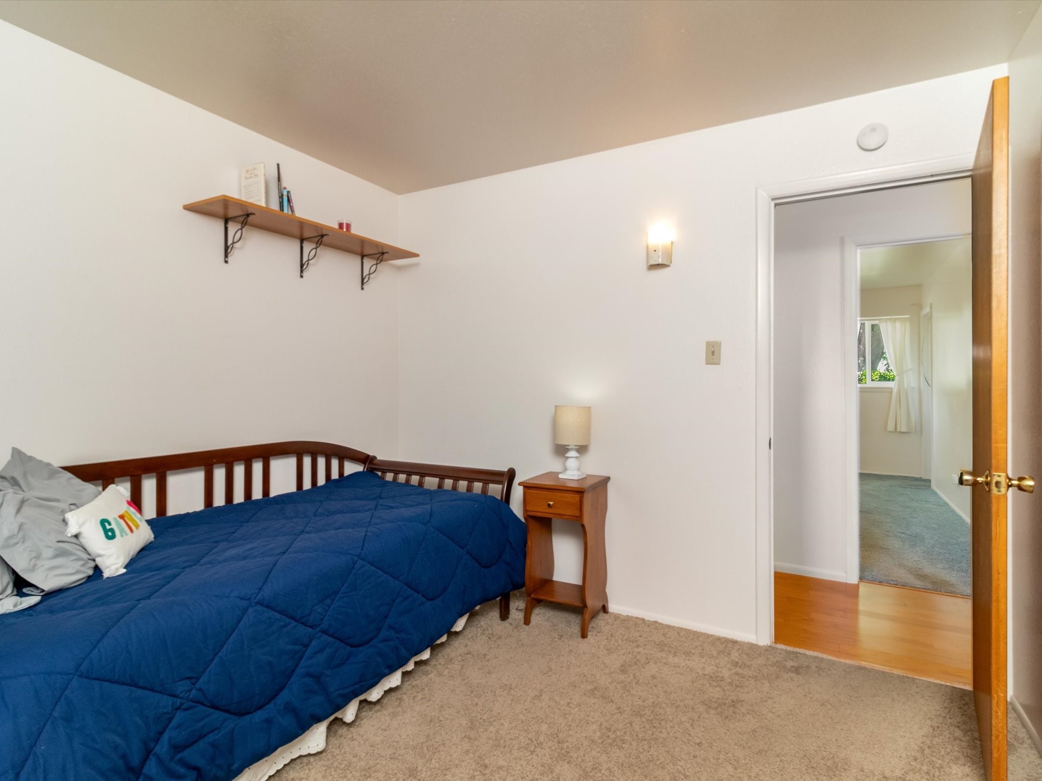 Bedroom with a blue-quilted single bed, wooden bedside table, and open doorway to the hall.