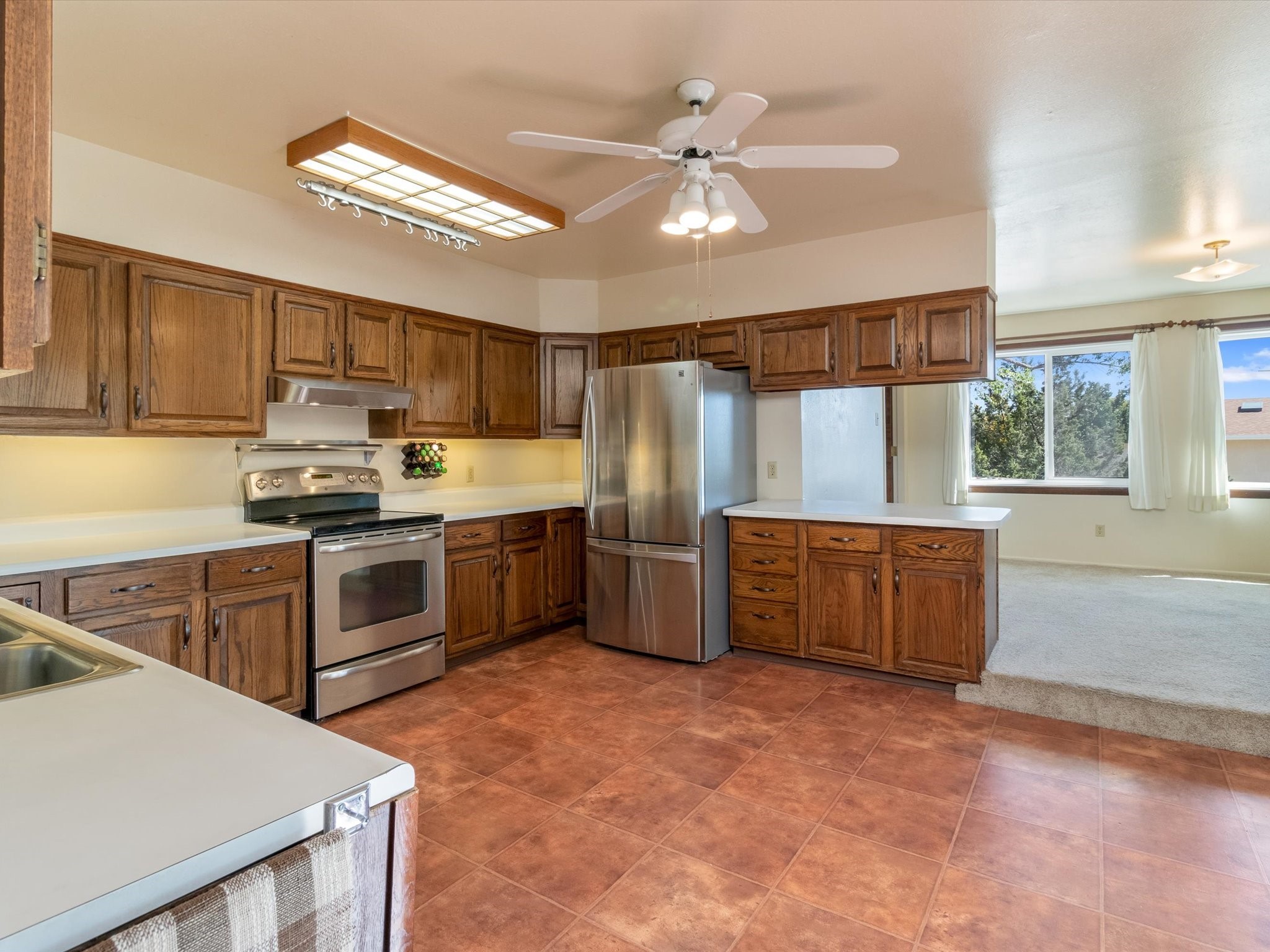 Kitchen with wood cabinets, stainless steel appliances, tile flooring, and a window overlooking trees.