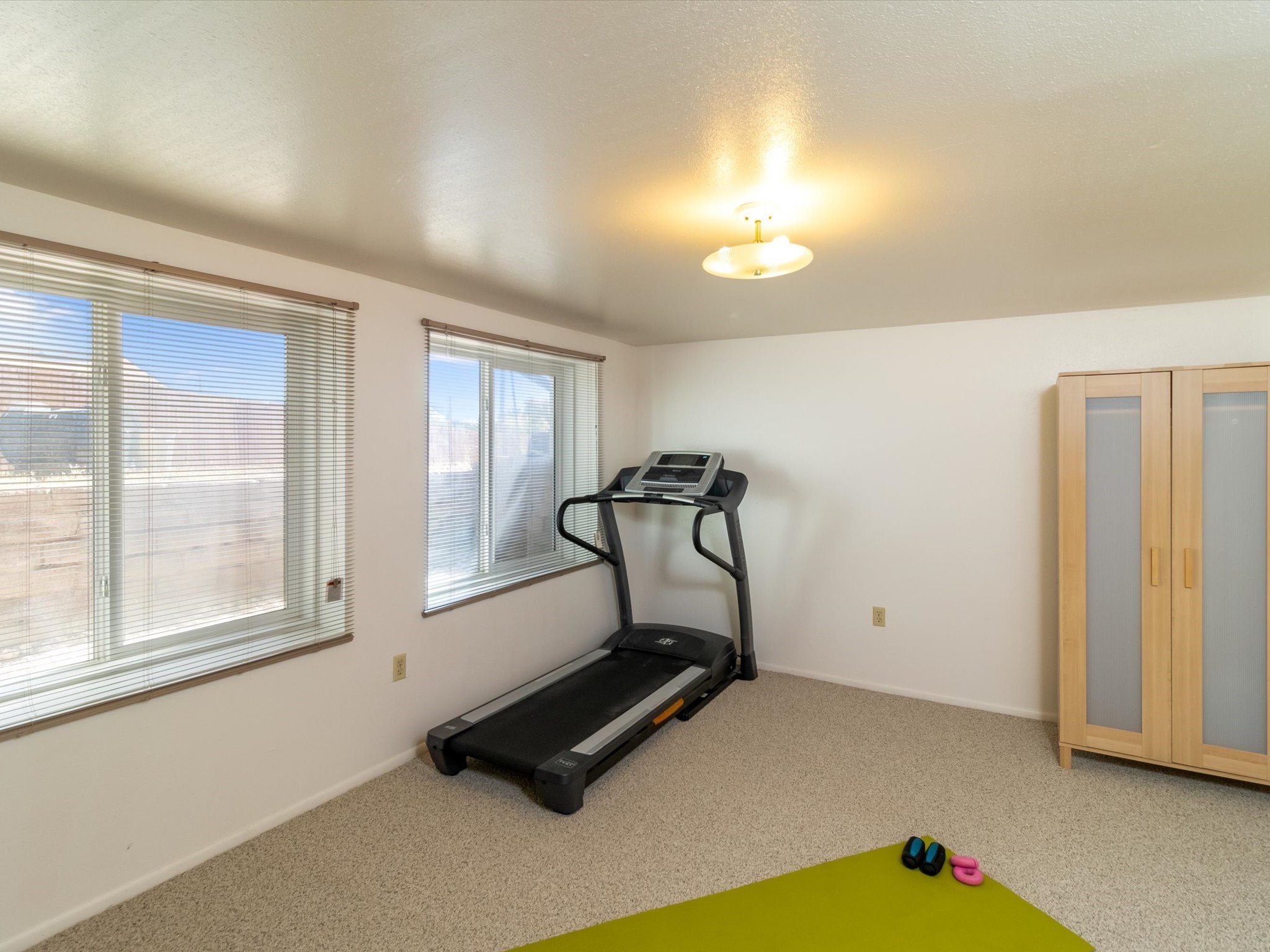 Exercise room with a treadmill, green yoga mat, and weights beside two windows.