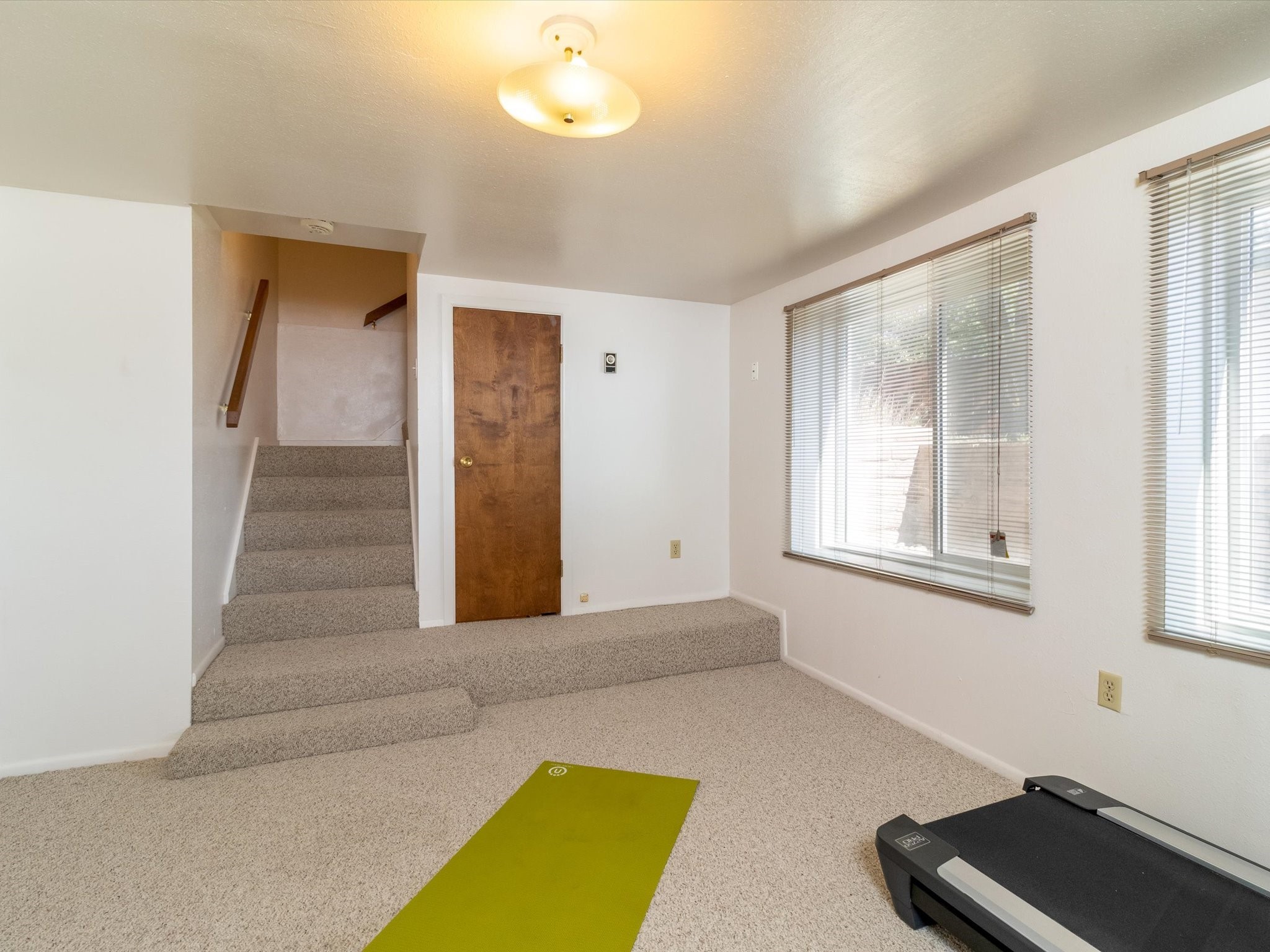 Carpeted room with stairs, two large windows, a green exercise mat, and a treadmill.