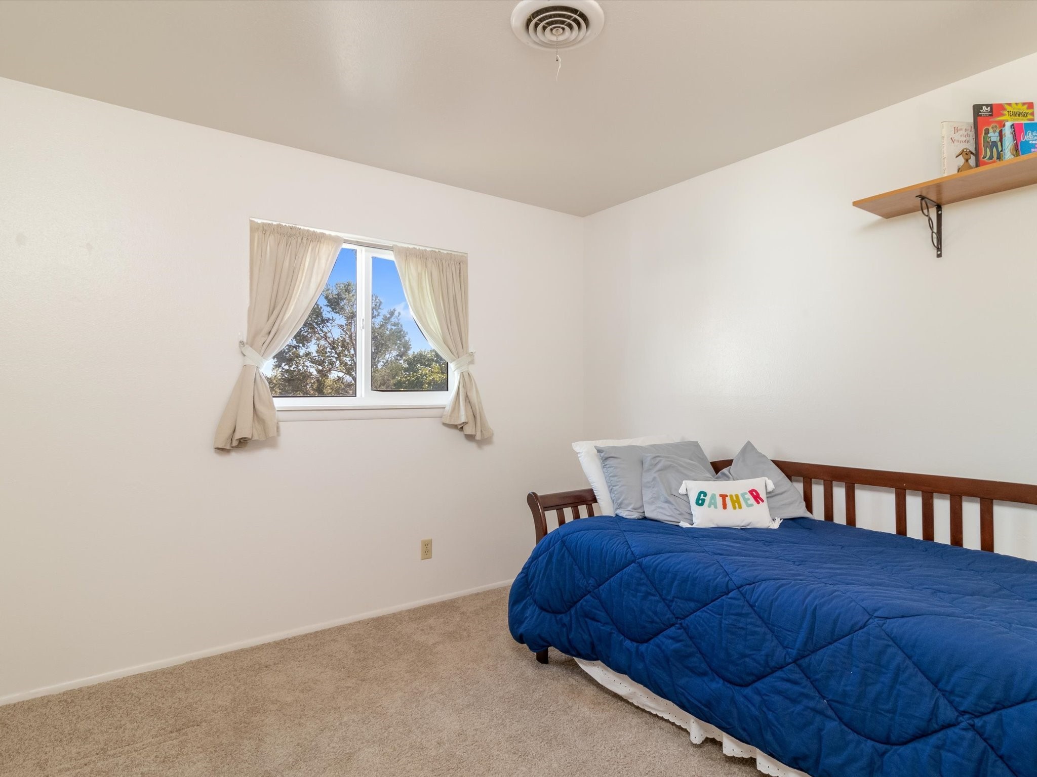 Bedroom with a blue quilted bed, curtained window, and a pillow reading “GATHER.”