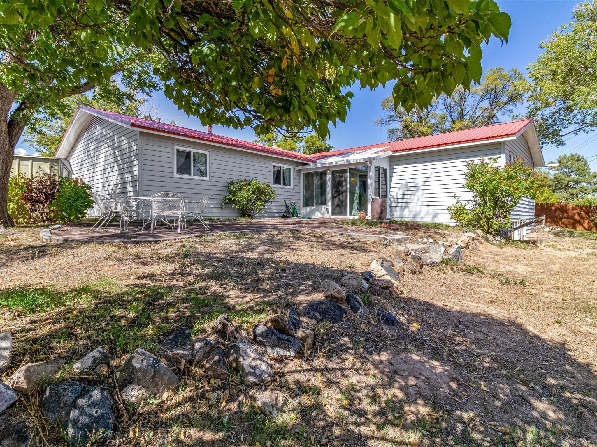 Rear of a gray-sided home with a red roof, shaded patio, and rocky yard.