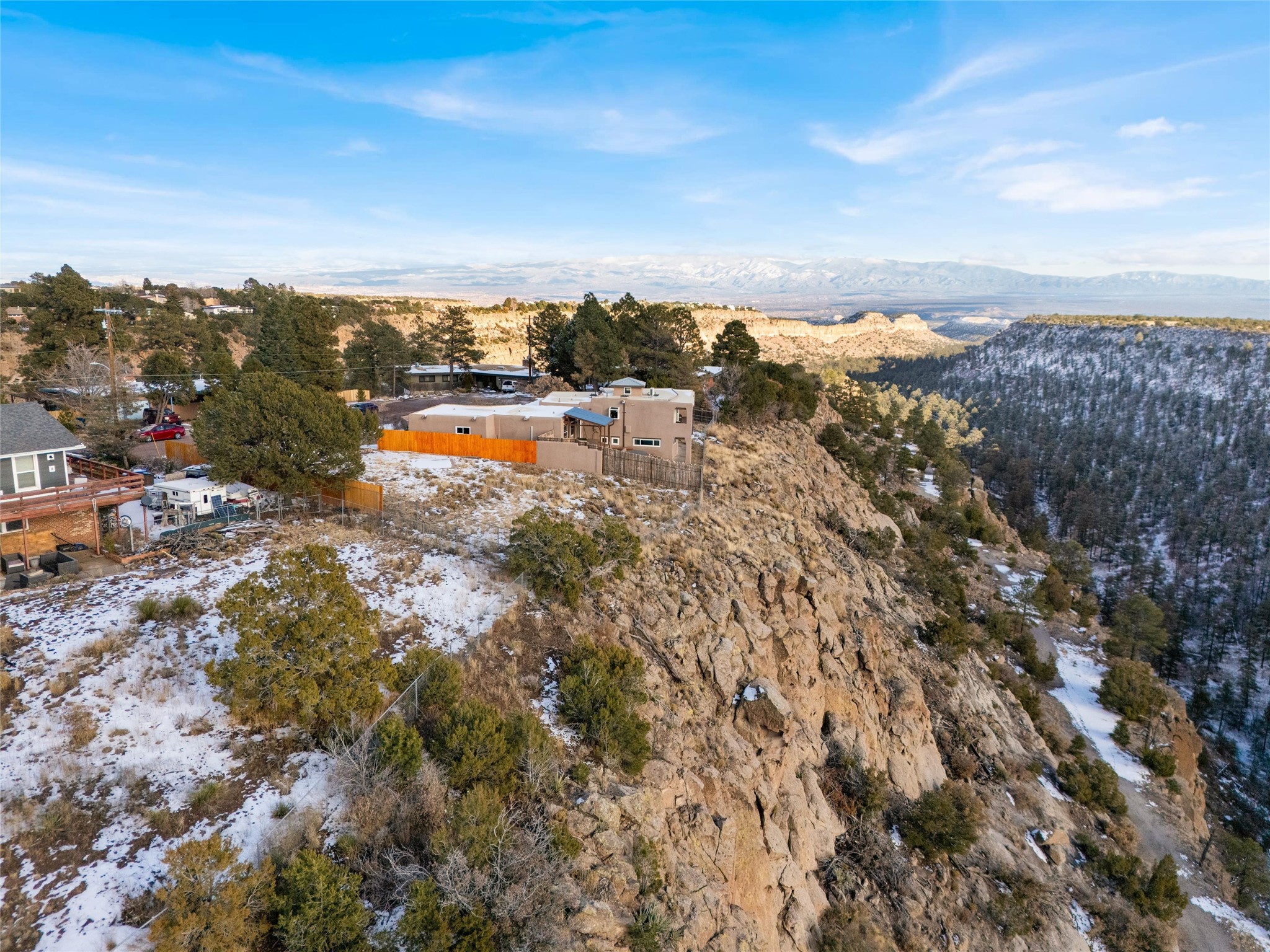 Homes on a rocky, snow-dusted ridge above a wooded canyon, with distant mountains.