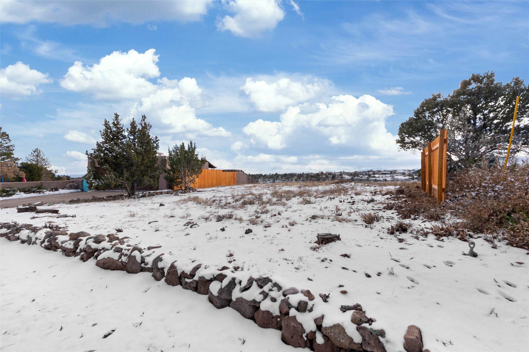 Snow-covered vacant lot with a low stone wall and wooden fencing.