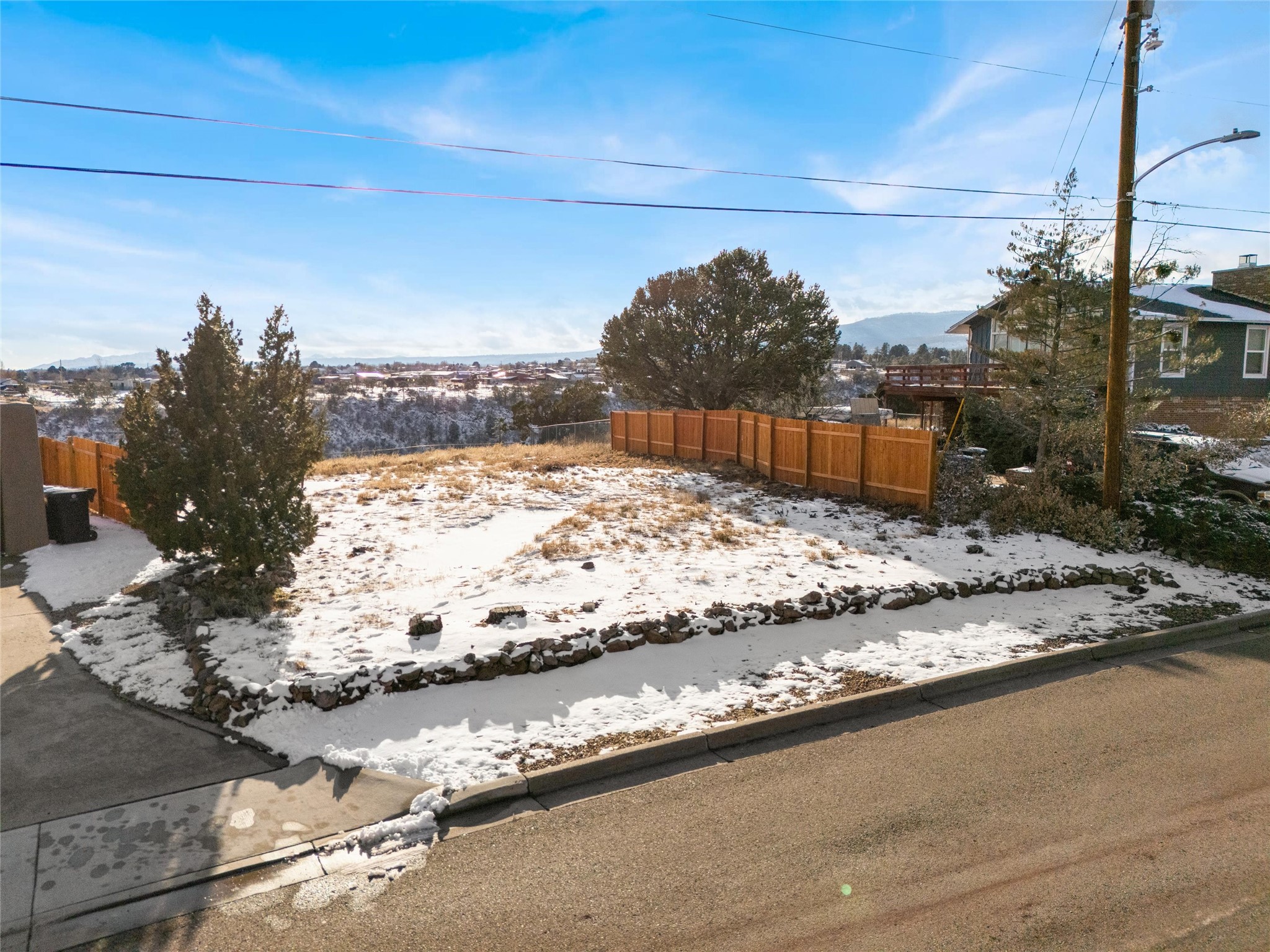 Snow-dusted fenced yard with a wooden fence, stone border, and distant neighborhood views.