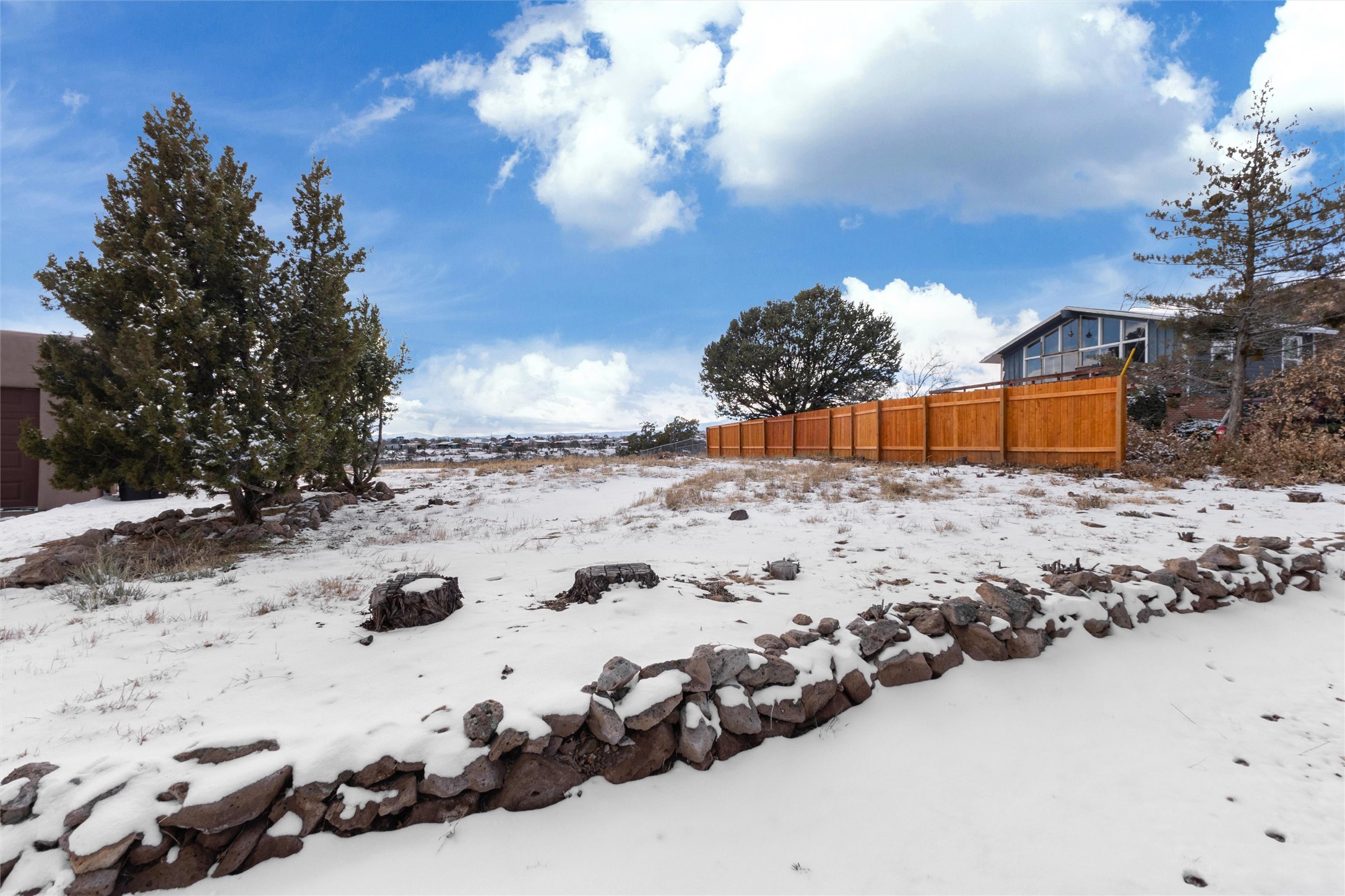 Snow-covered vacant lot with a stone wall, tree stumps, and a long wooden fence.