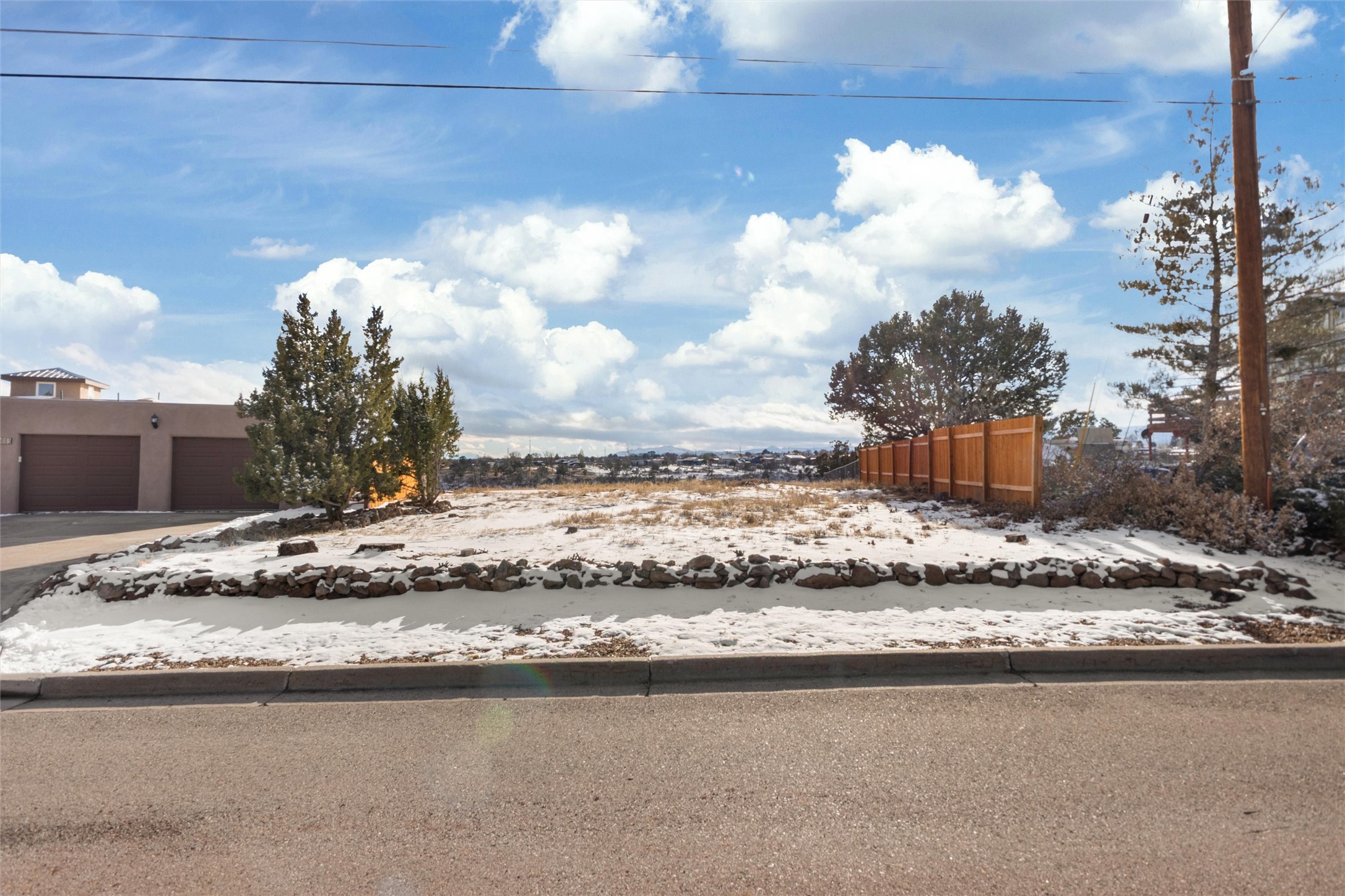 Snow-covered vacant lot bordered by stones, with a wooden fence and neighboring garage.
