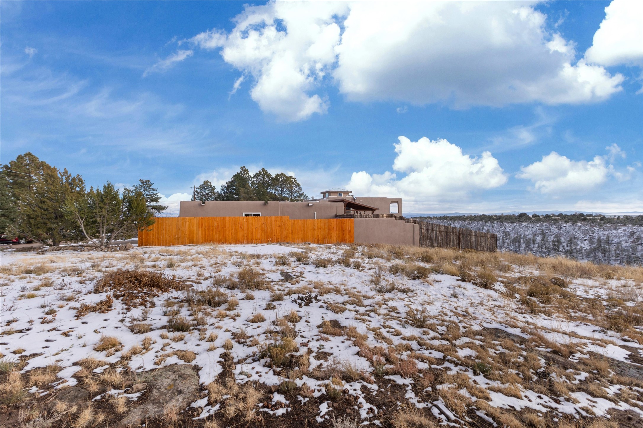 Adobe-style home behind an orange fence on a snow-dusted lot, with wooded hills beyond.