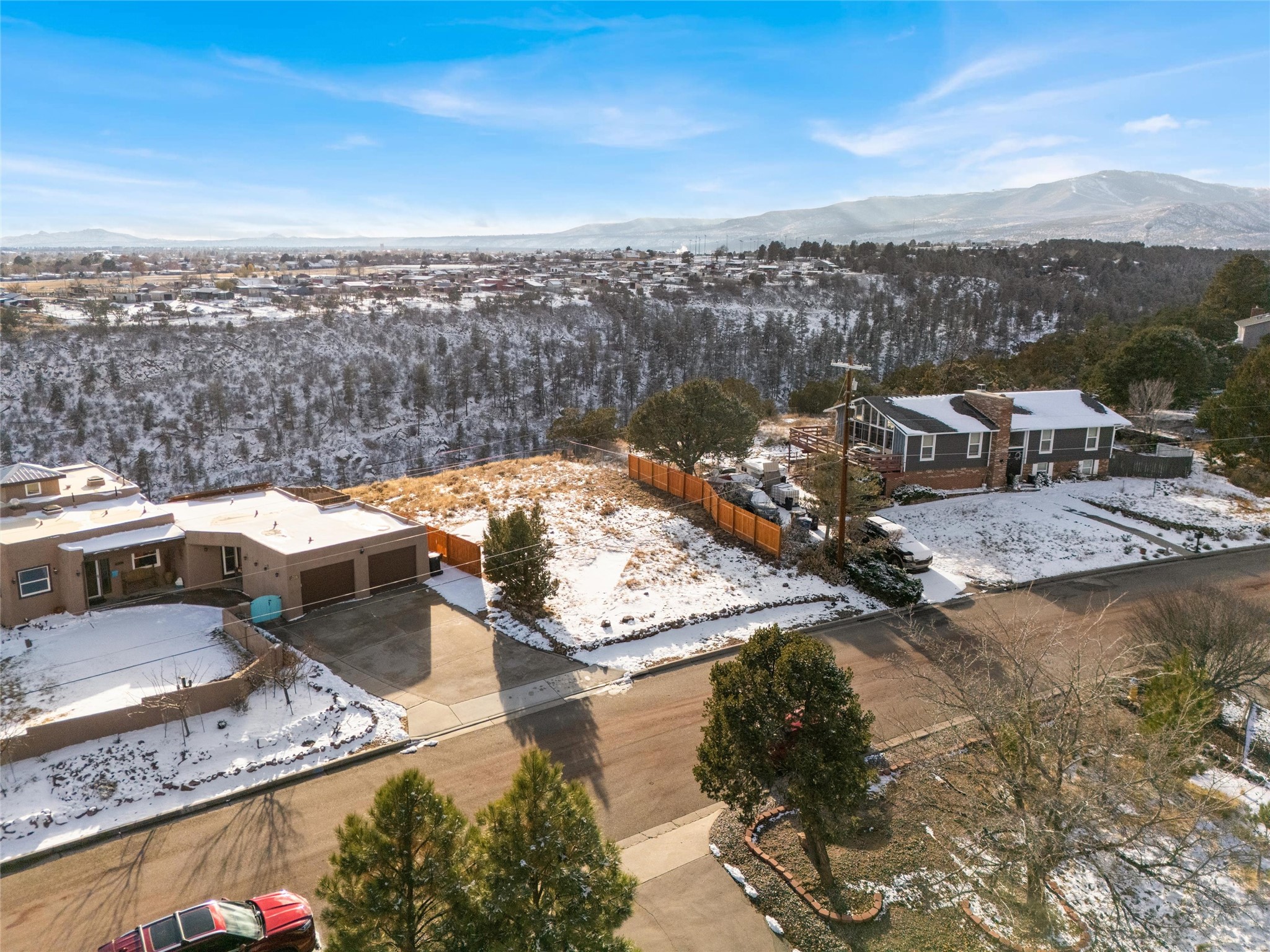 Aerial view of snow-covered homes and a vacant lot, with a wooded ravine and mountains beyond.