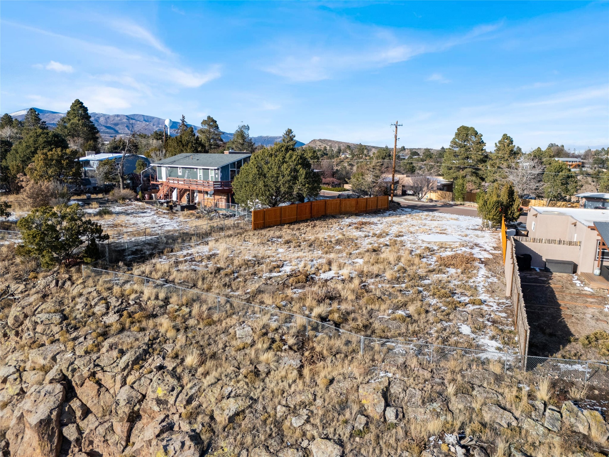 Rocky vacant lot with patchy snow, neighboring homes, and mountain views.