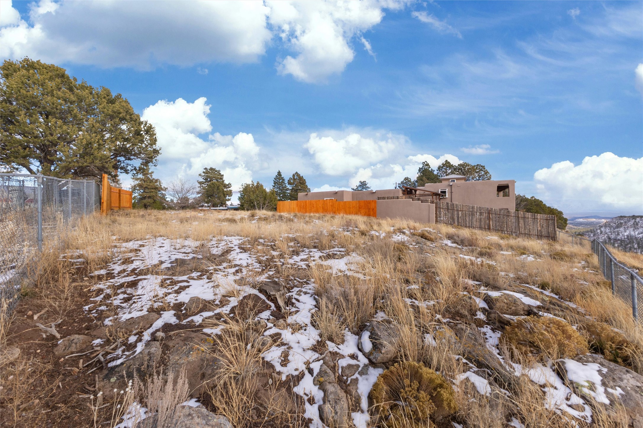 Modern stucco home above a rocky, snow-patched slope, with fenced boundaries and a distant valley.