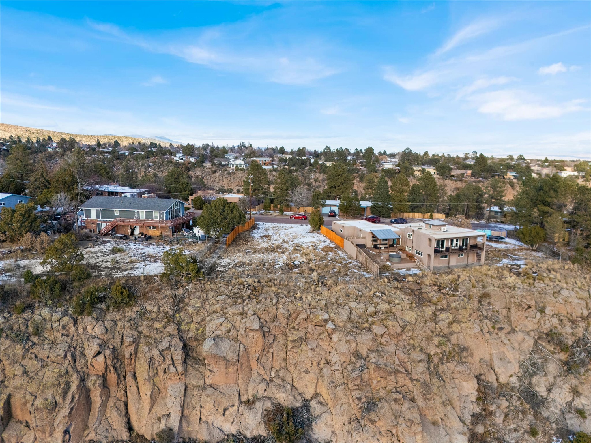Aerial view of a tan two-story home beside a rocky bluff, with snow-dusted ground and neighborhood homes beyond.