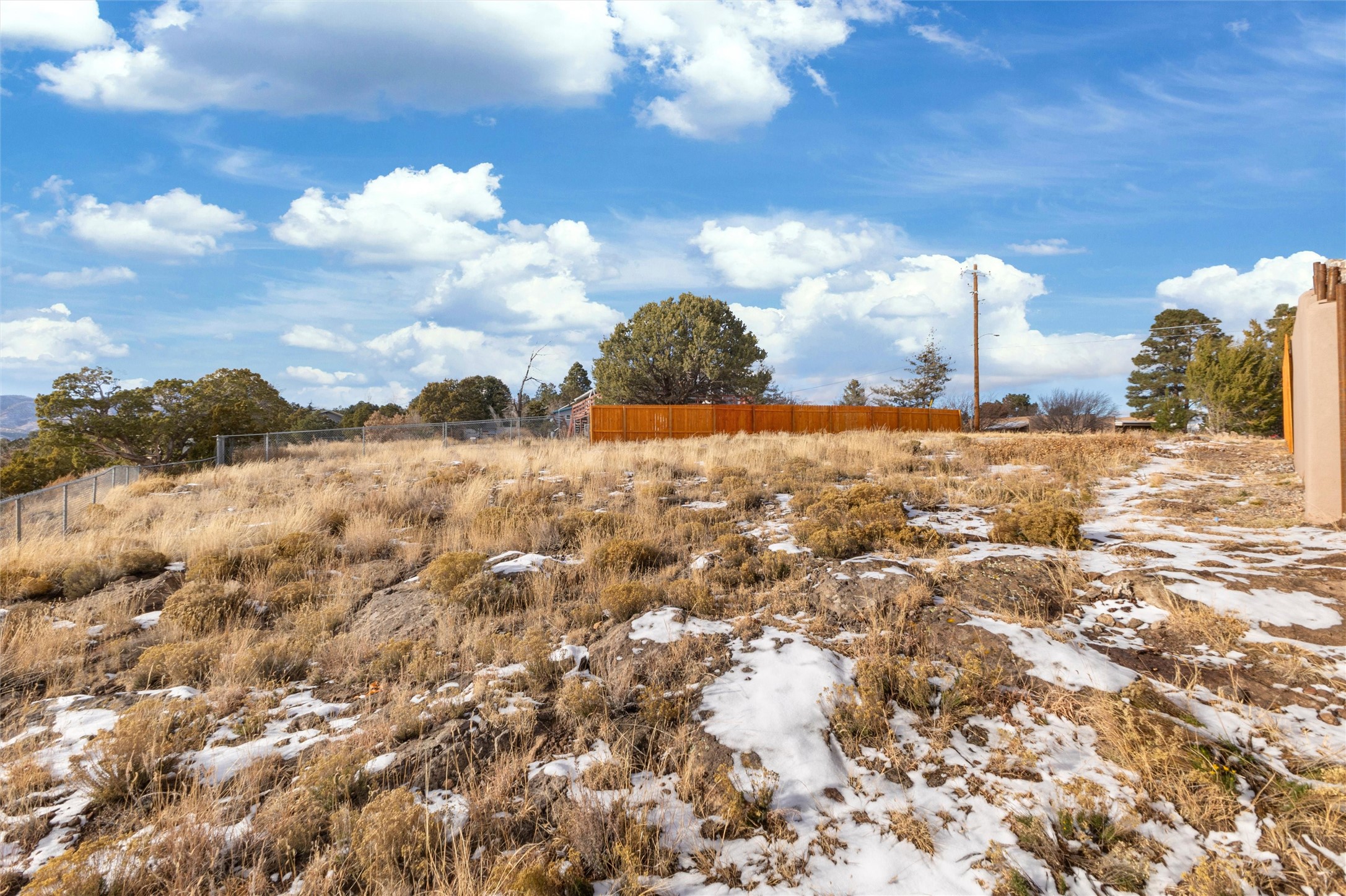 Snow-dusted vacant lot with dry grasses, rocky ground, and fencing beside a stucco home.