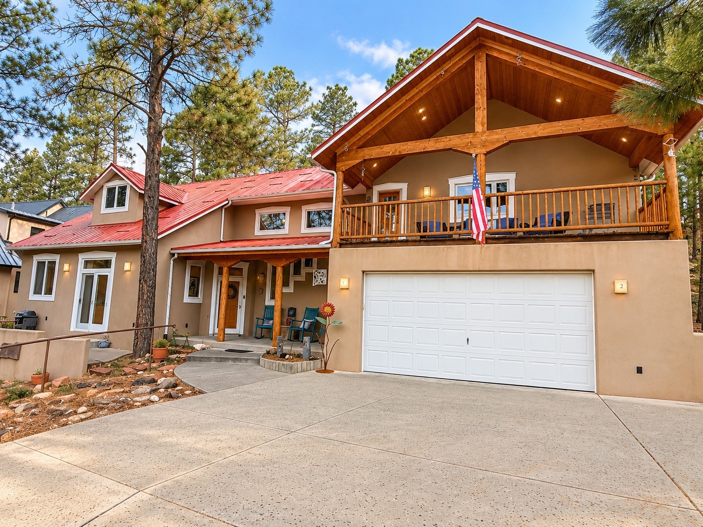 Two-story tan home with red metal roof, timber balcony, and wide concrete driveway among pine trees.