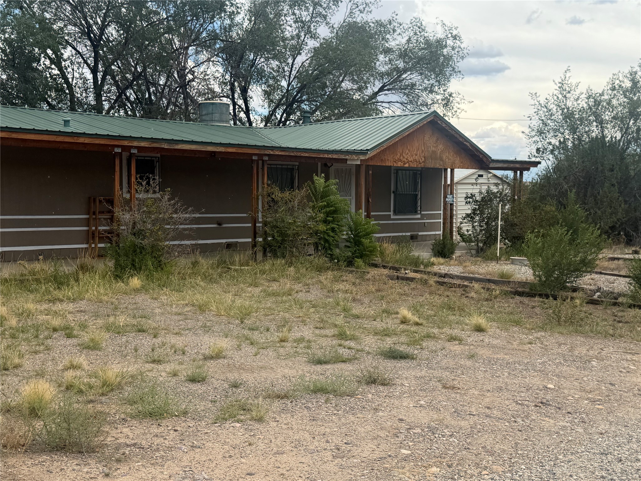 Single-story home with a green metal roof, covered porch, and sparse vegetation across the gravel yard.