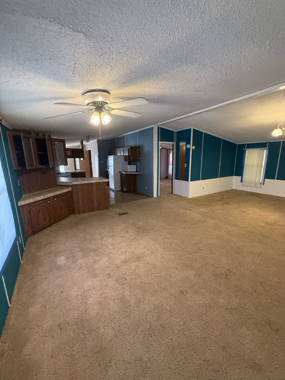 Vacant living room with tan carpet, teal walls, and an adjoining kitchen with dark wood cabinets.