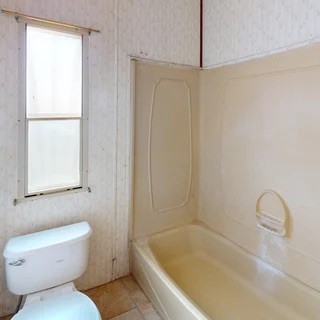 Empty bathroom with a beige bathtub, white toilet, and window.