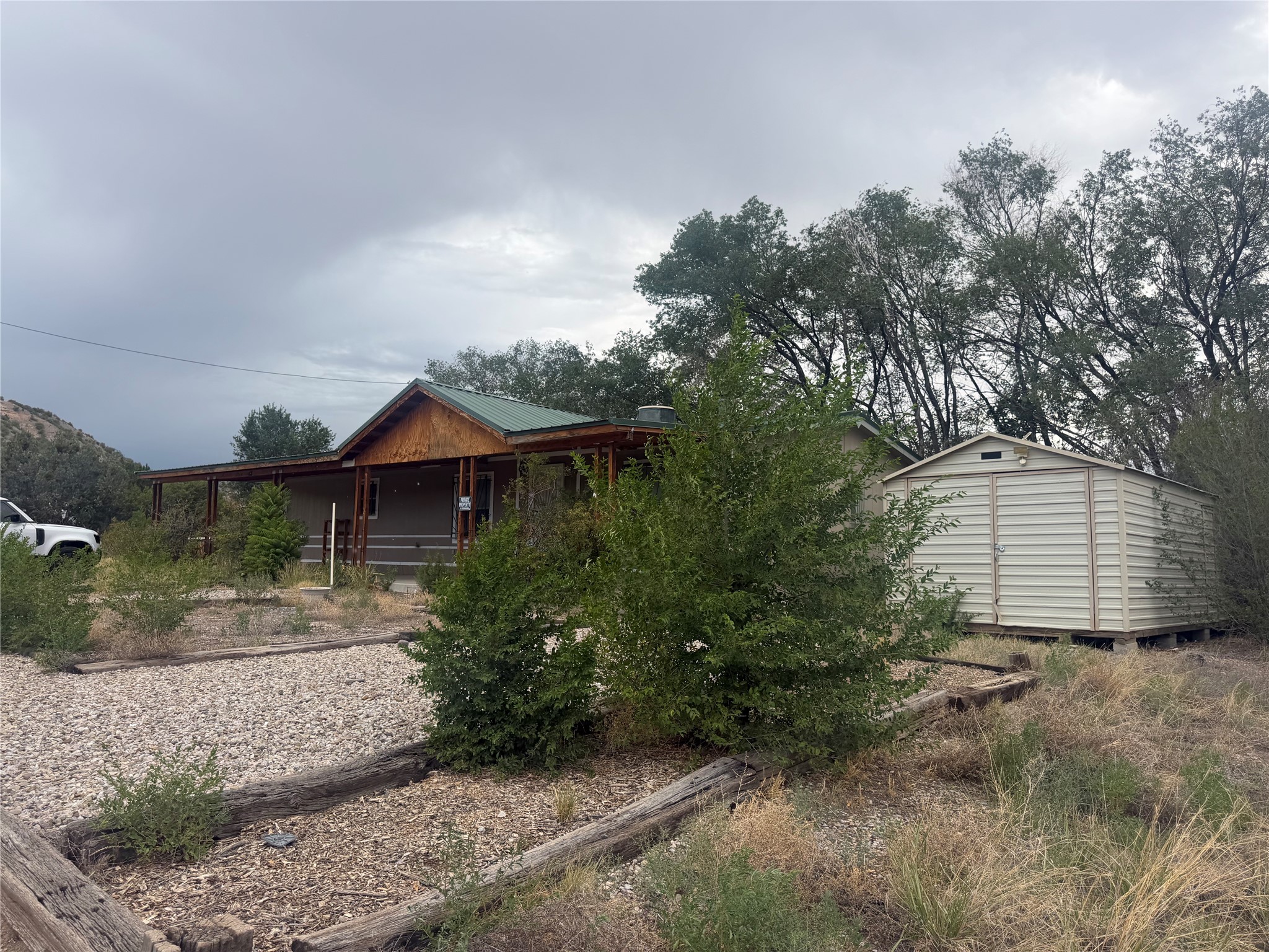 Single-story home with a green metal roof and covered porch, partly screened by shrubs beside a detached shed.