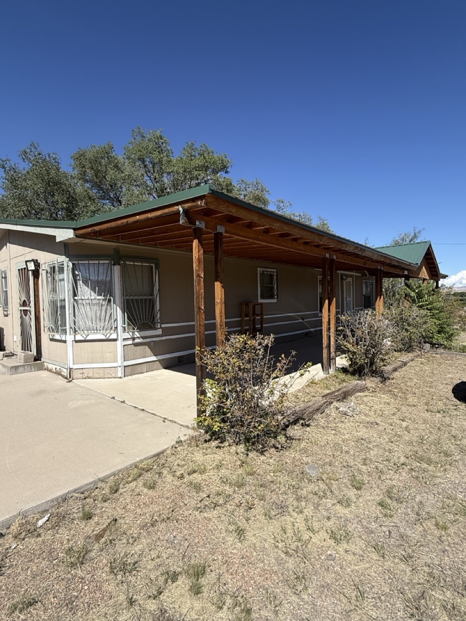 Single-story home with a long wooden-post porch, green roof, and dry, sparsely planted yard.