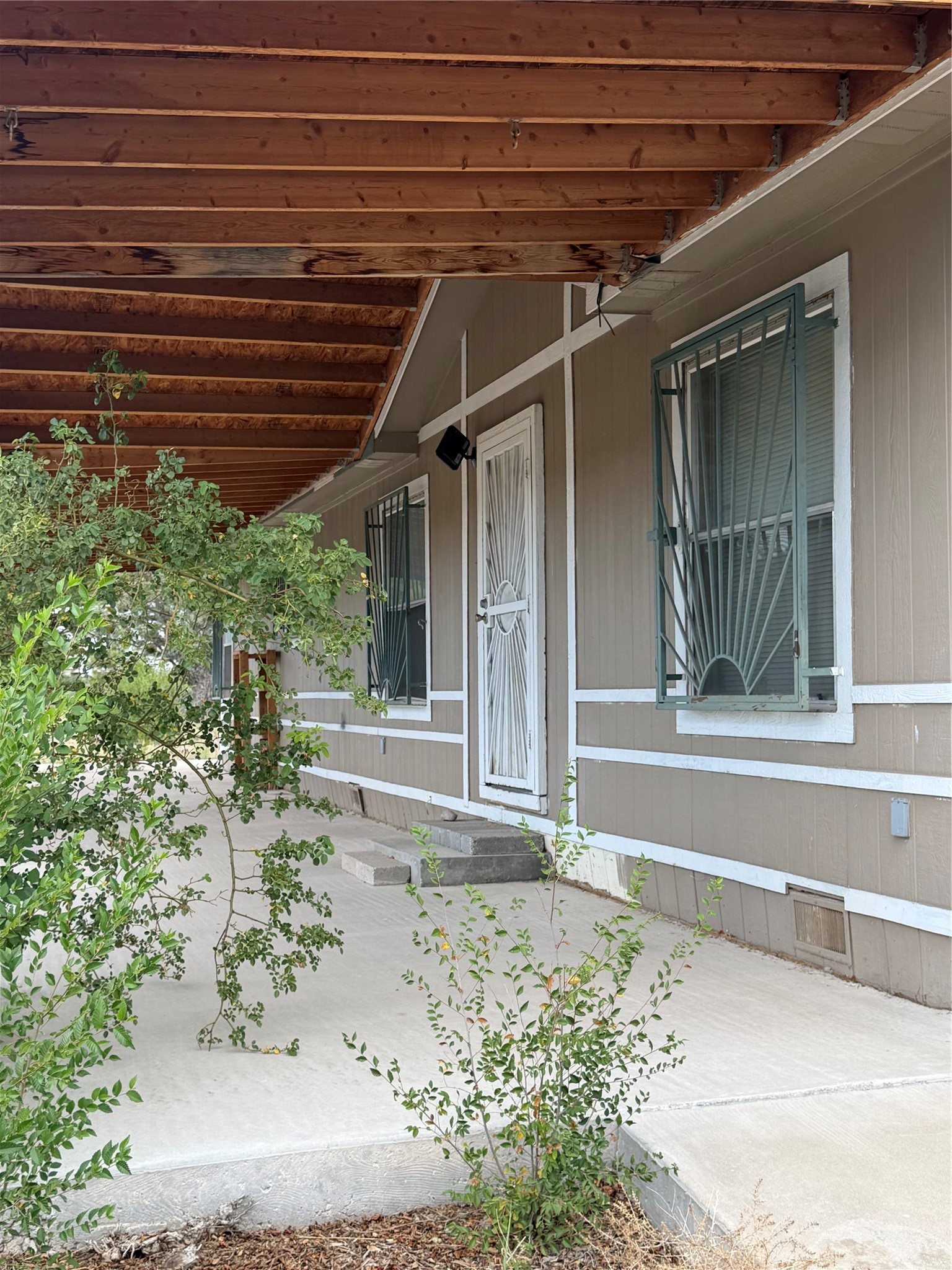 Home’s covered porch with a white security door, barred windows, and shrubs along the concrete walkway.
