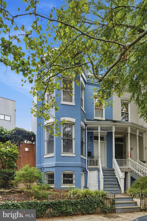 Blue townhouse with a covered porch, white railings, and front steps beneath leafy tree branches.