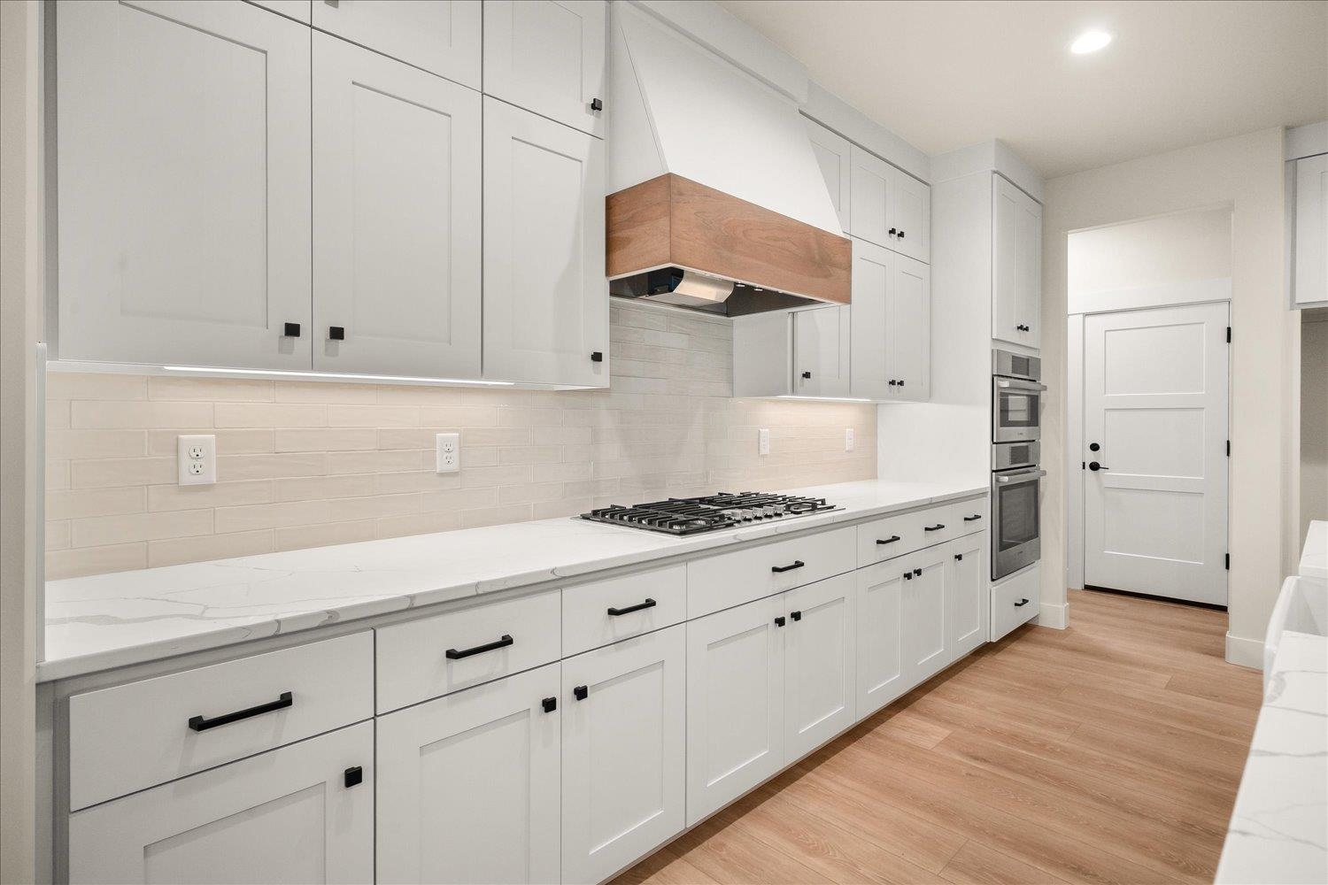 Modern white kitchen with a gas cooktop, wood-trimmed range hood, and warm wood flooring.