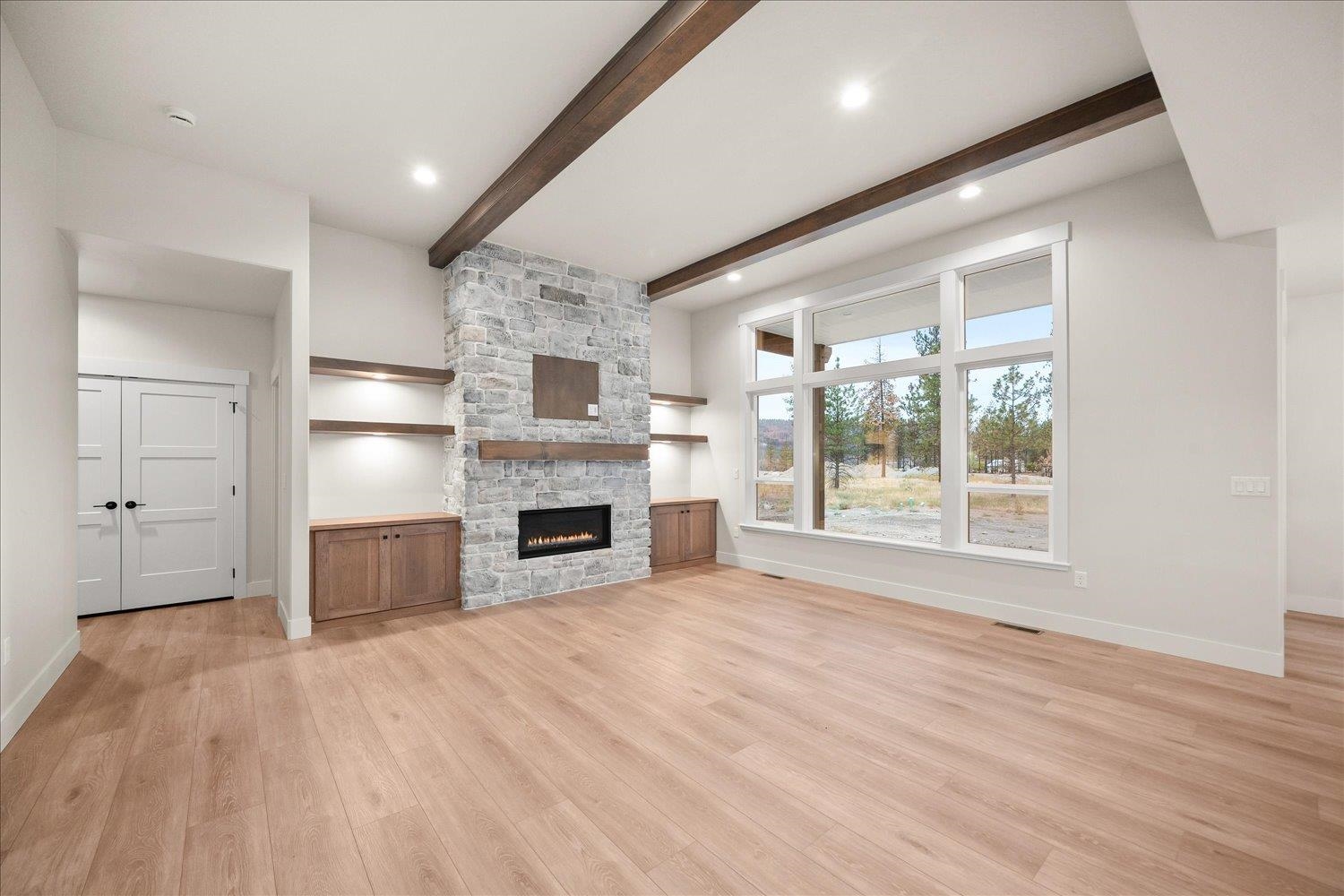 Empty living room with stone fireplace, dark ceiling beams, built-in cabinets, and a large picture window.