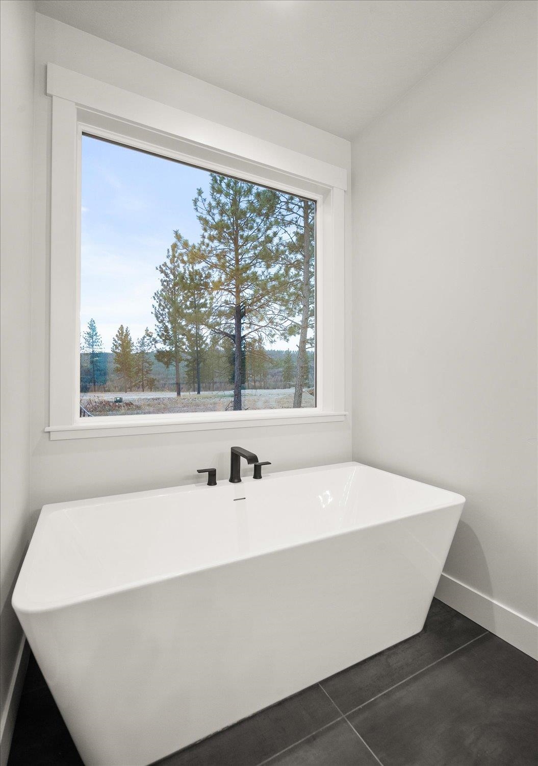 Modern bathroom with a freestanding tub beneath a large window overlooking pine trees.