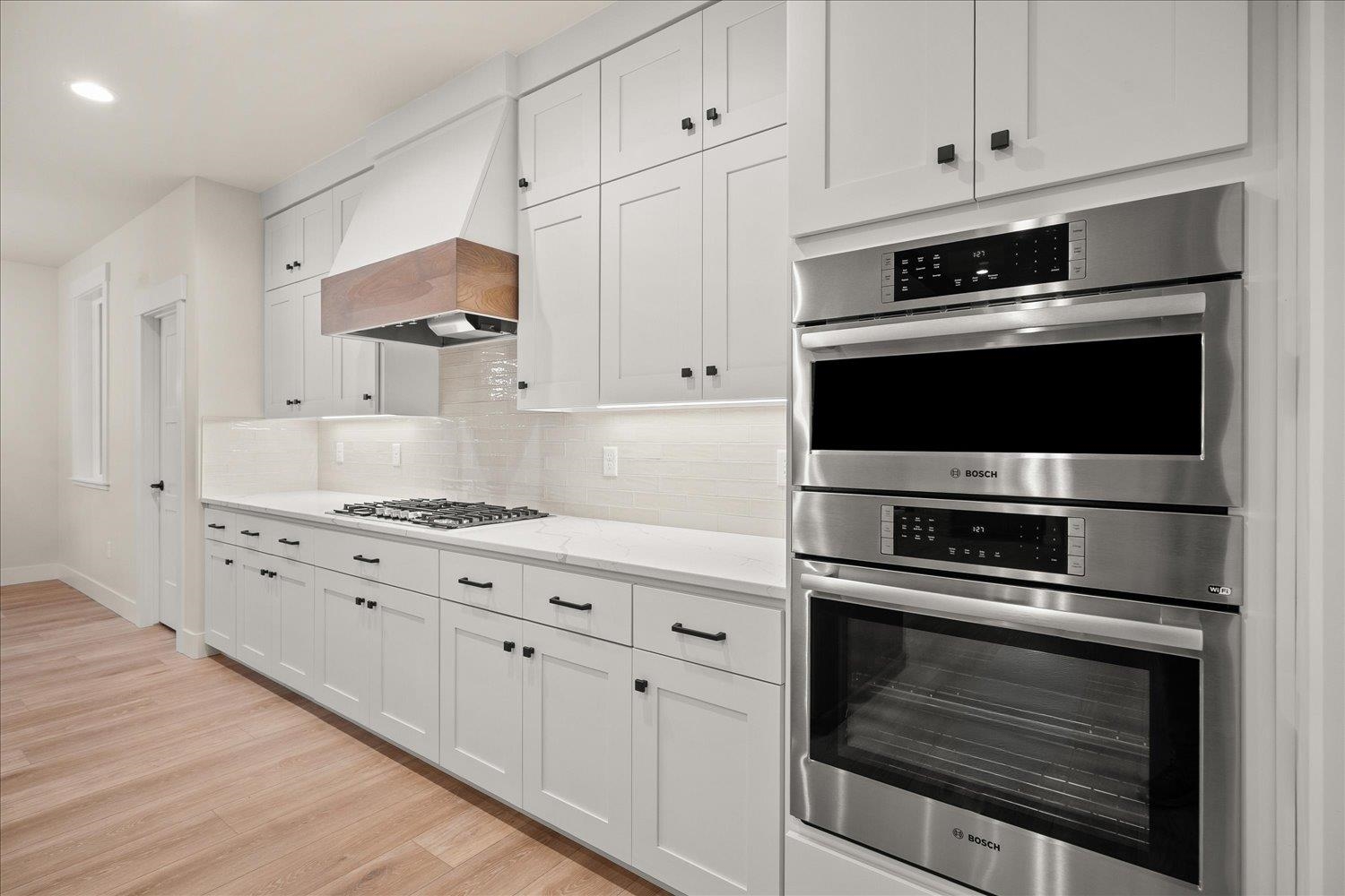 Bright white kitchen with gas cooktop, wood-trimmed hood, and stainless double wall ovens.