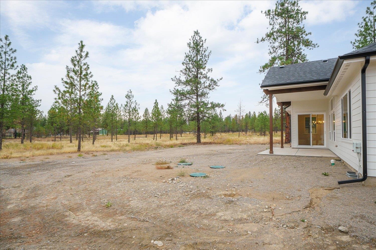 White house with a covered patio beside an open gravel yard and pine trees.