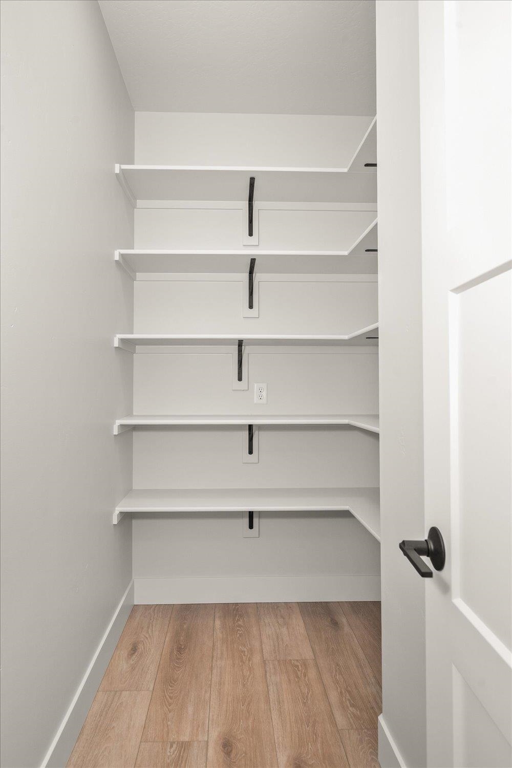 Empty walk-in closet with white shelves, black brackets, and wood-look flooring.
