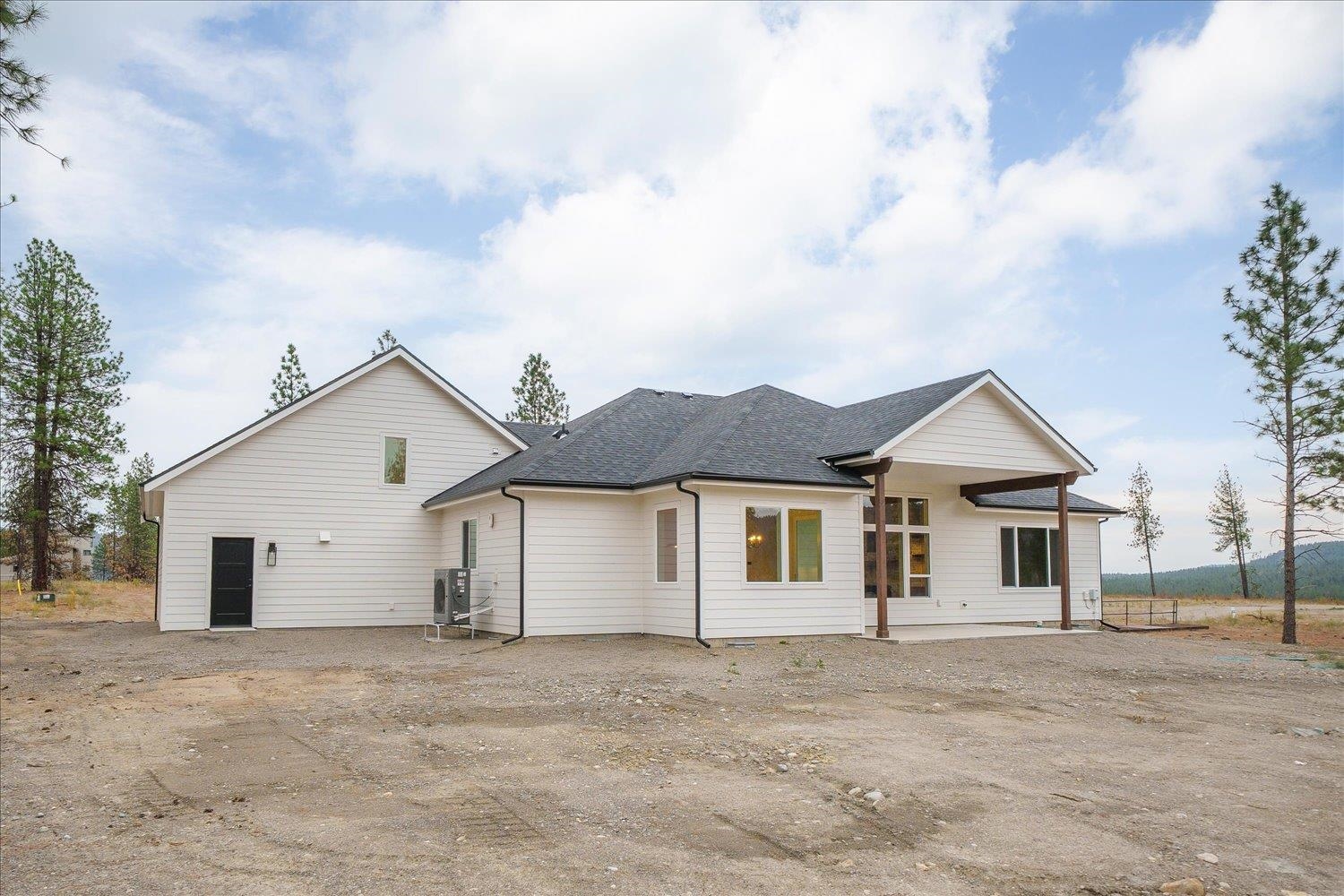White single-story home with a covered patio and gravel yard, backed by evergreen trees and wooded hills.