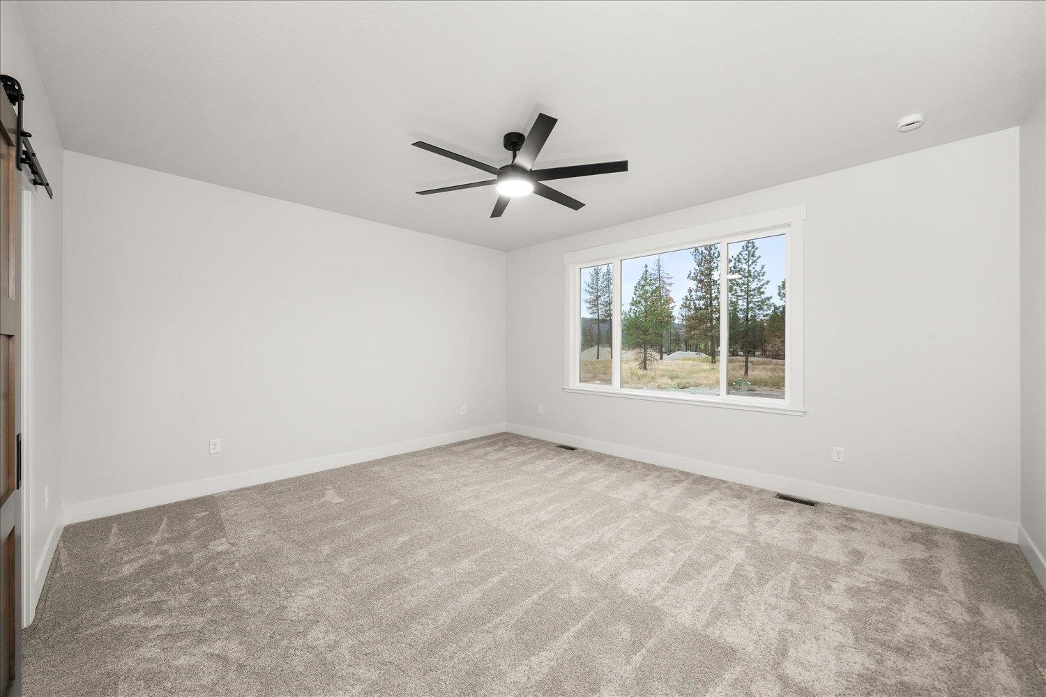 Empty bedroom with gray carpet, a black ceiling fan, and a large window overlooking pine trees.