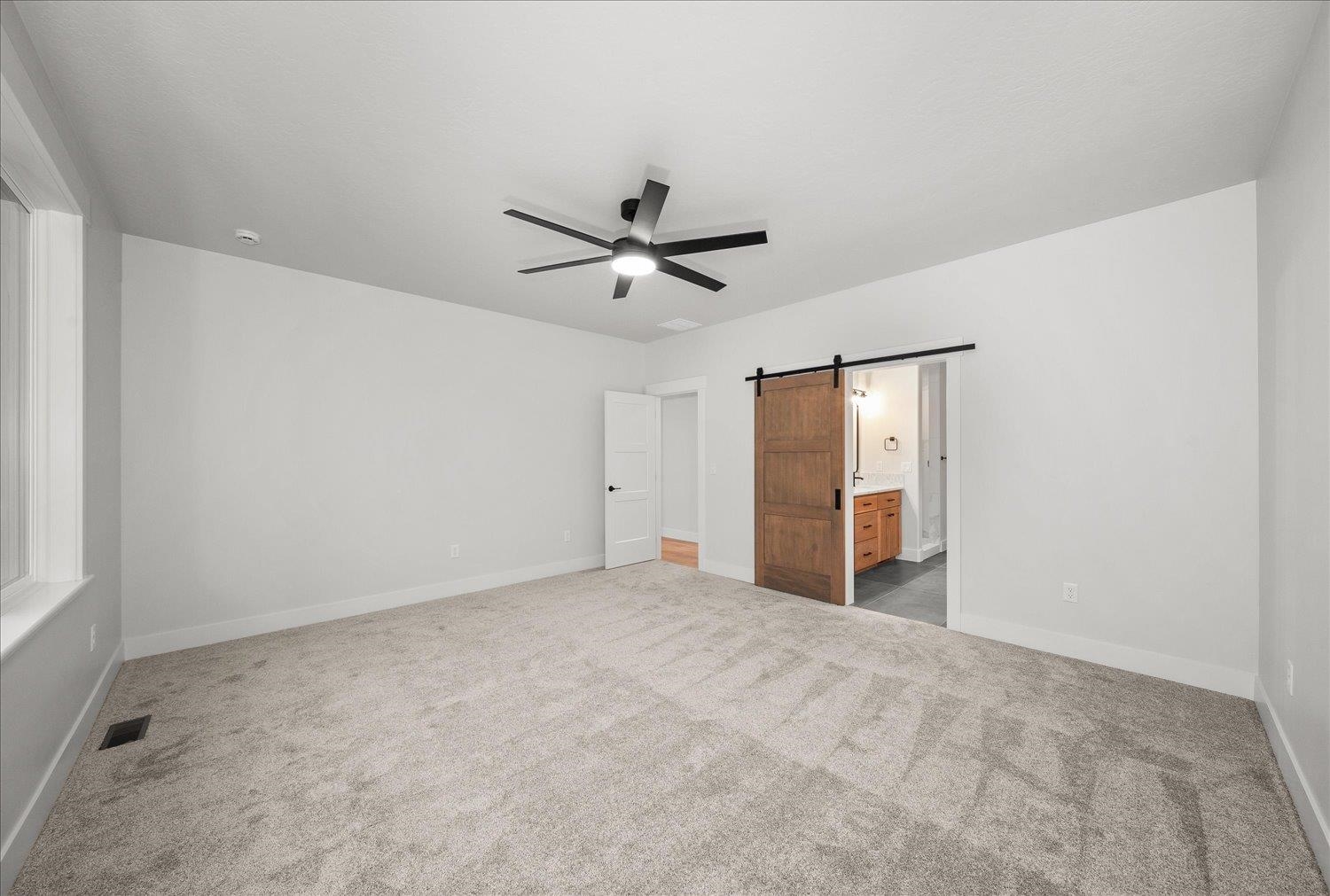 Empty carpeted bedroom with a black ceiling fan and a wood barn door opening toward the bathroom.