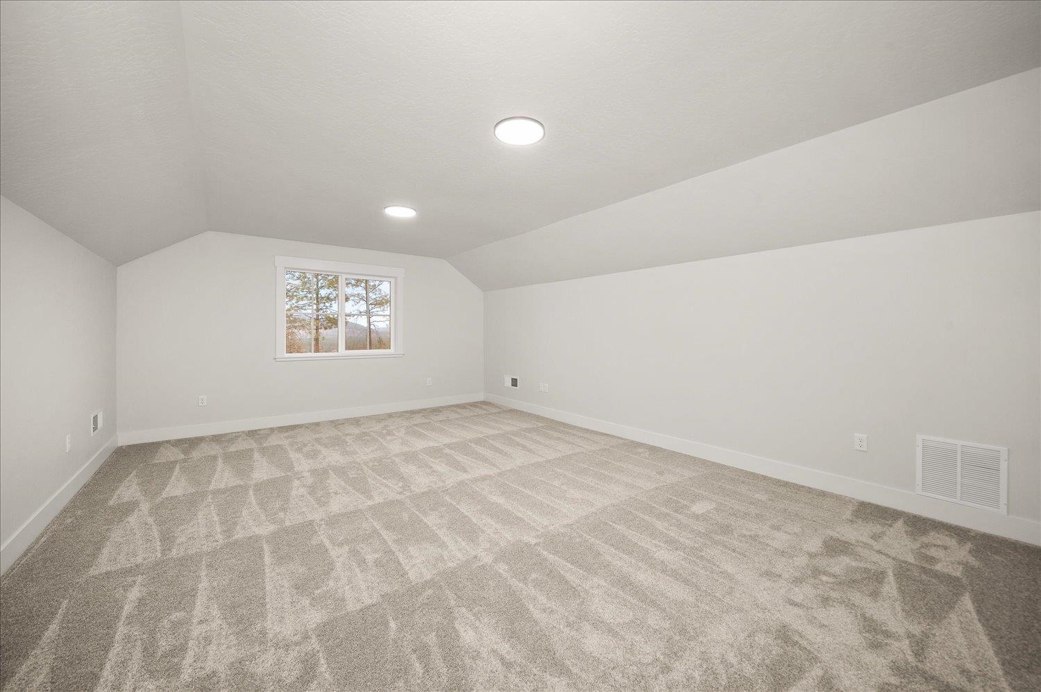 Empty carpeted room with a high angled ceiling, recessed lights, and a window overlooking trees.