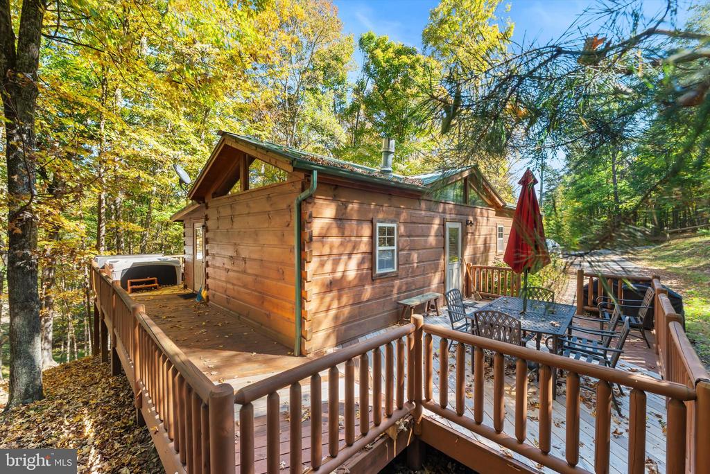 Wood-sided cabin with a raised deck, outdoor dining furniture, and a hot tub among autumn trees.