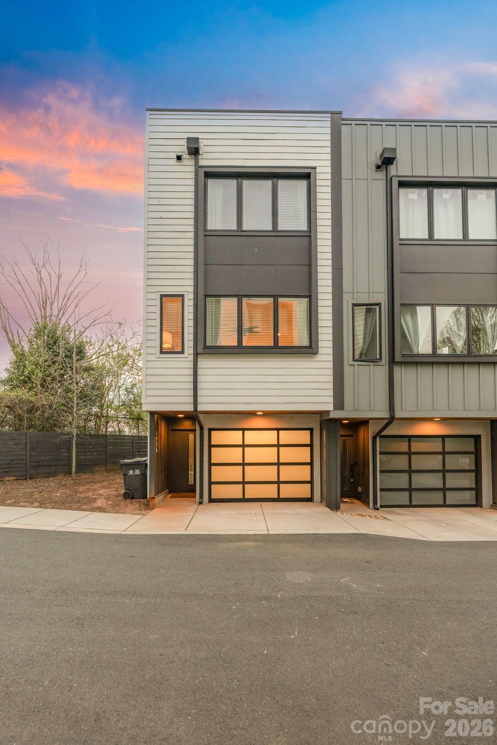 Modern attached townhomes with white and gray siding and illuminated garage doors beneath a pink dusk sky.