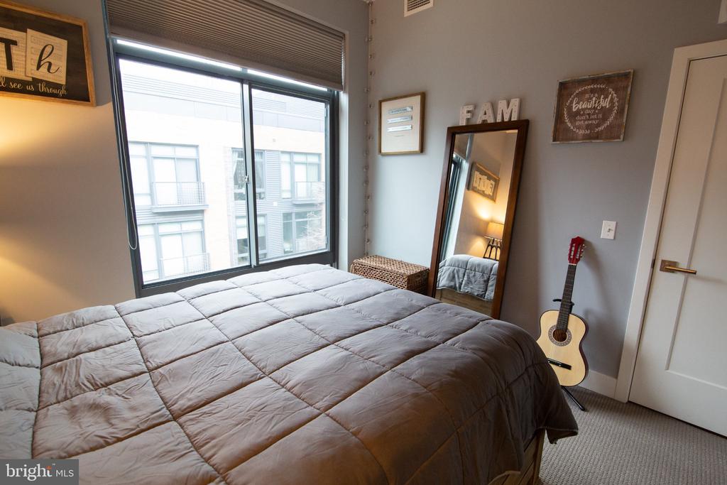 Furnished bedroom with a large gray-quilted bed, tall window, leaning mirror, and guitar.