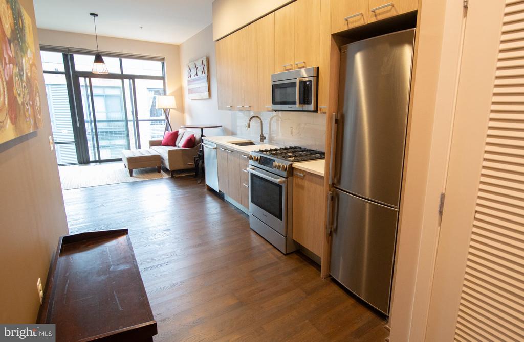 Condo kitchen with light wood cabinets and stainless steel appliances opening to a windowed living area.