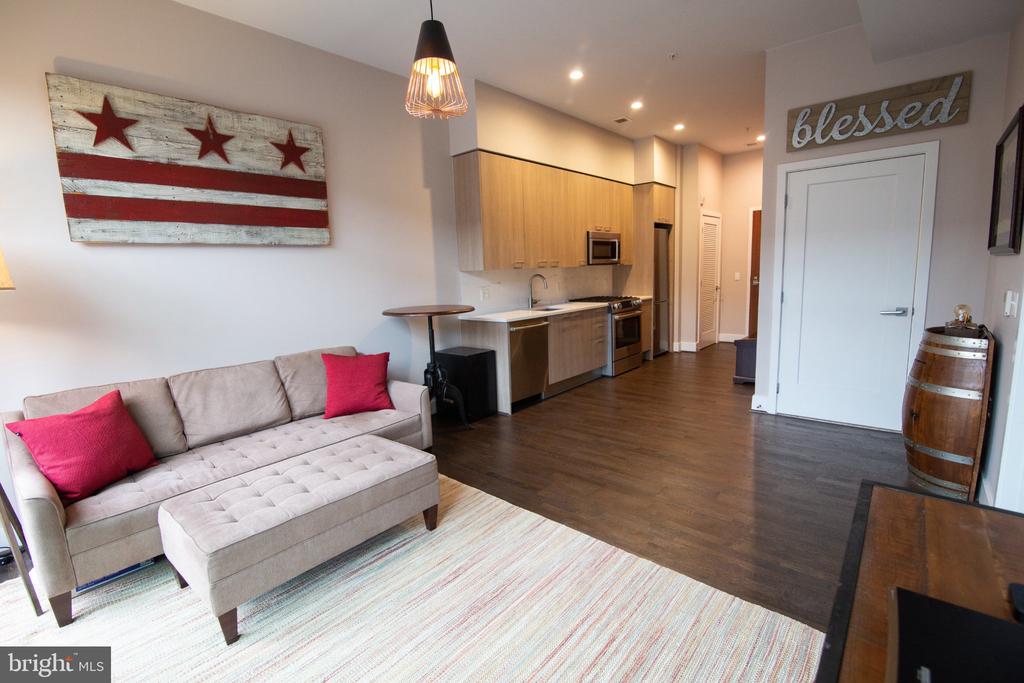 Open-plan condo living room and kitchen with a gray sectional, pink pillows, and light wood cabinets.