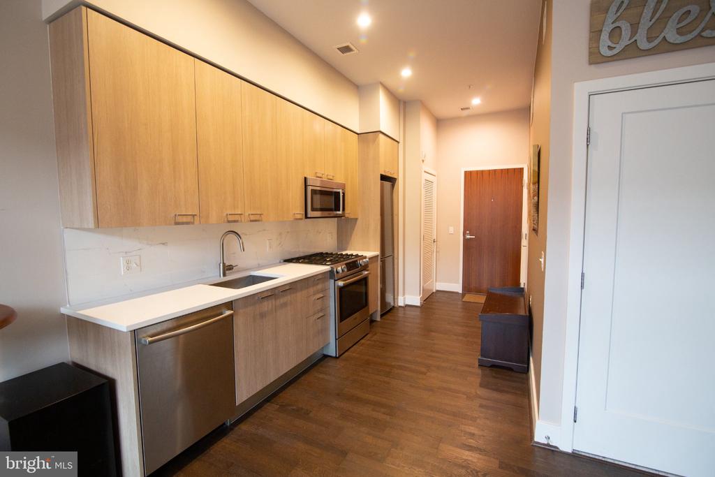 Modern galley kitchen with light wood cabinets, white counters, and stainless-steel appliances.